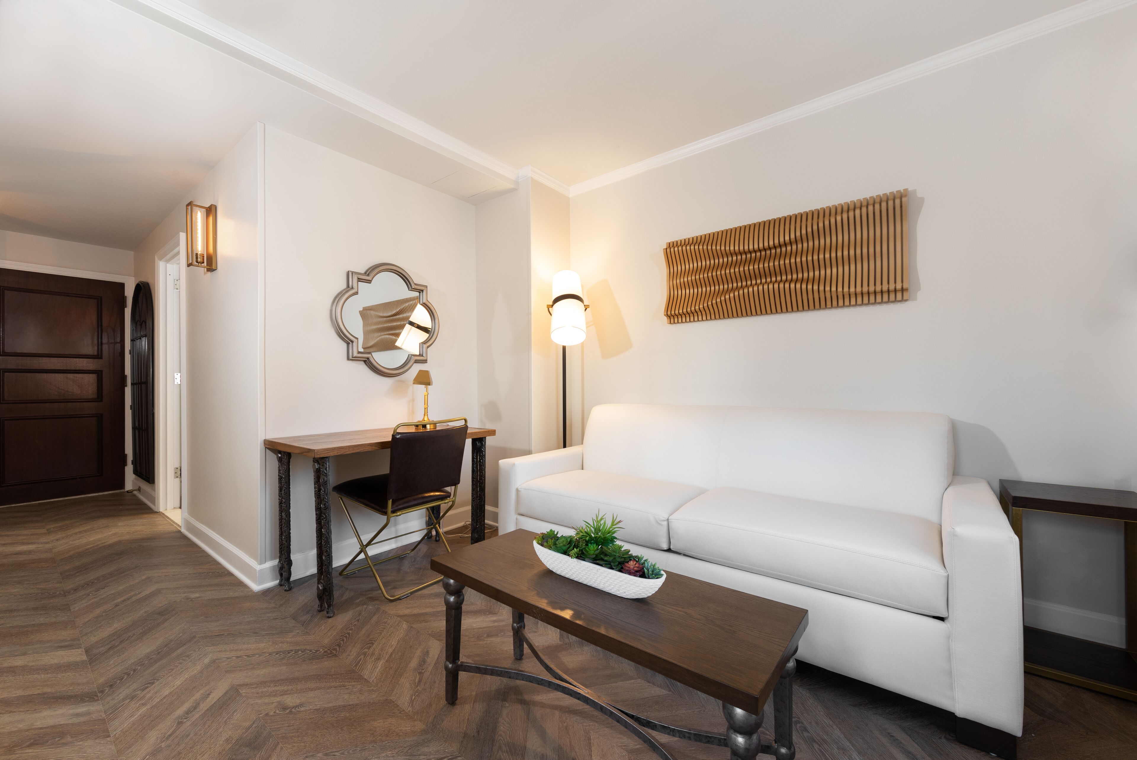 A modern living room with a white sofa, a wooden coffee table, a desk with a chair, and a lamp.