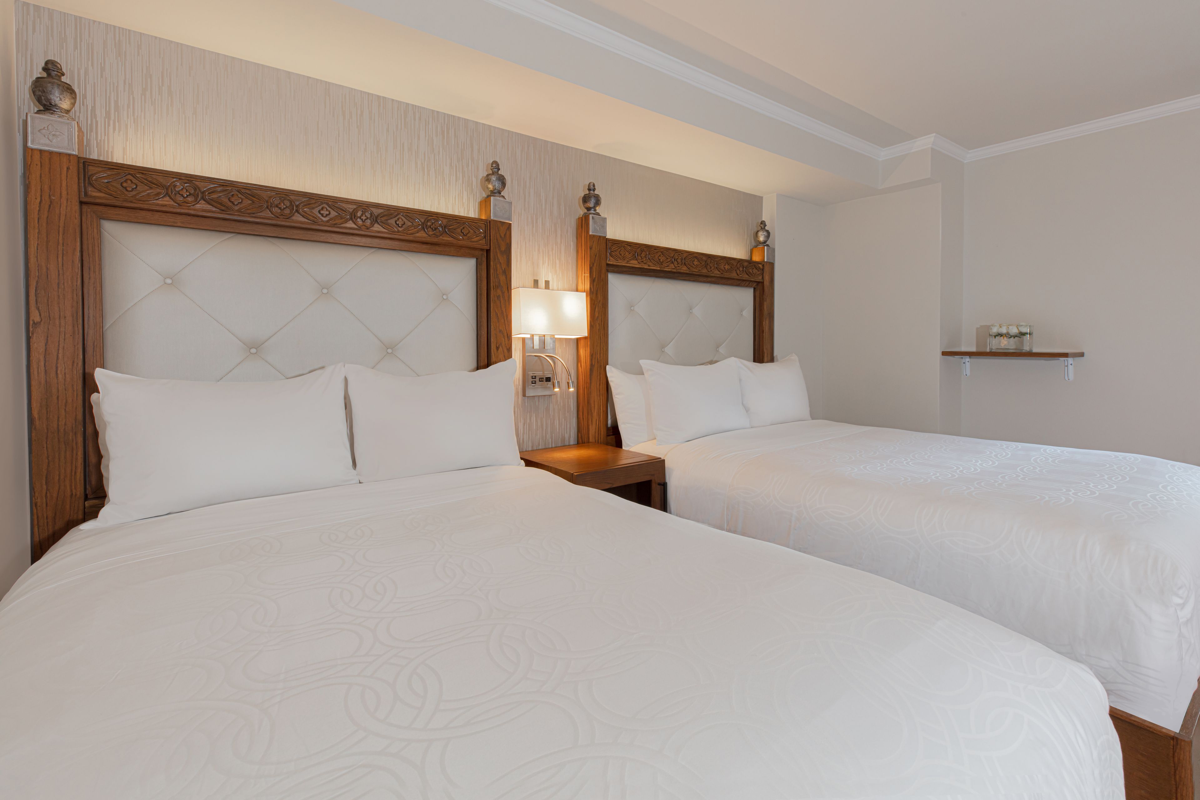 Habitación de hotel con dos camas tamaño queen, un cabecero de madera, una mesita de noche con lámpara y un estante sobre la cama.