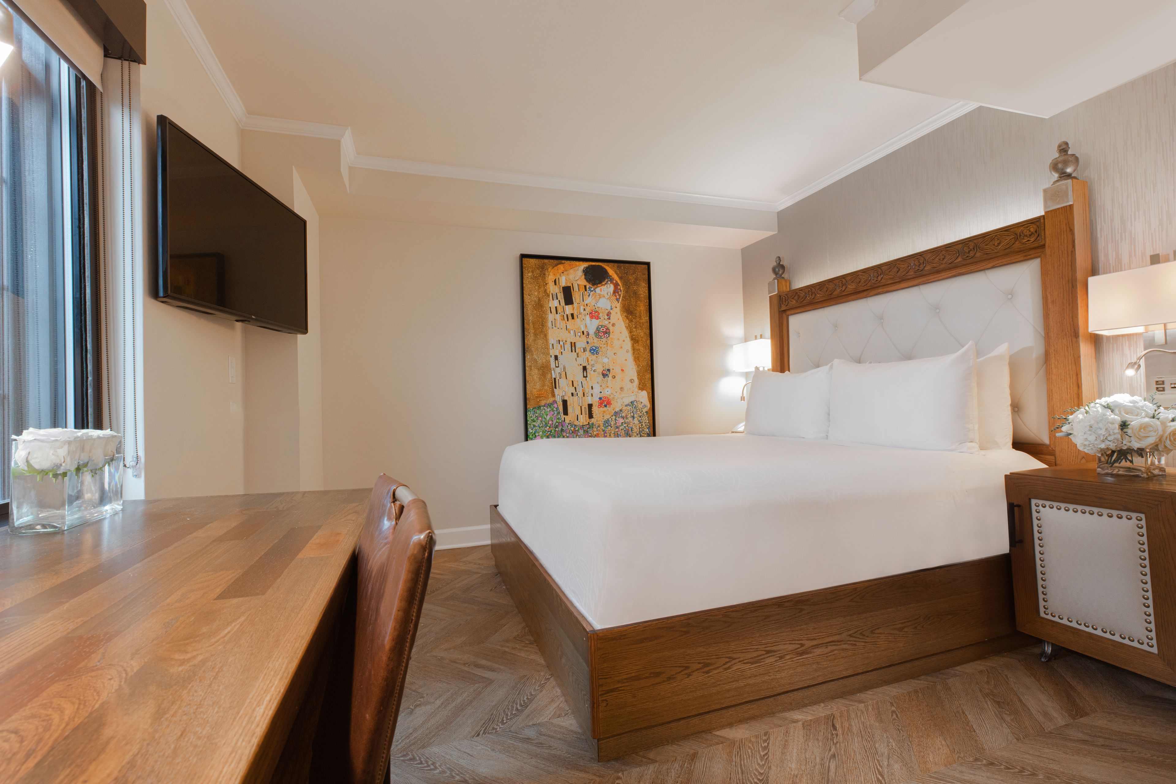Habitació d'hotel amb un llit de fusta, un televisor pla penjat a la paret, un quadre gran i una taula de nit amb una làmpada i flors.