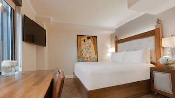 Habitació d'hotel amb un llit de fusta, un televisor pla penjat a la paret, un quadre gran i una taula de nit amb una làmpada i flors.