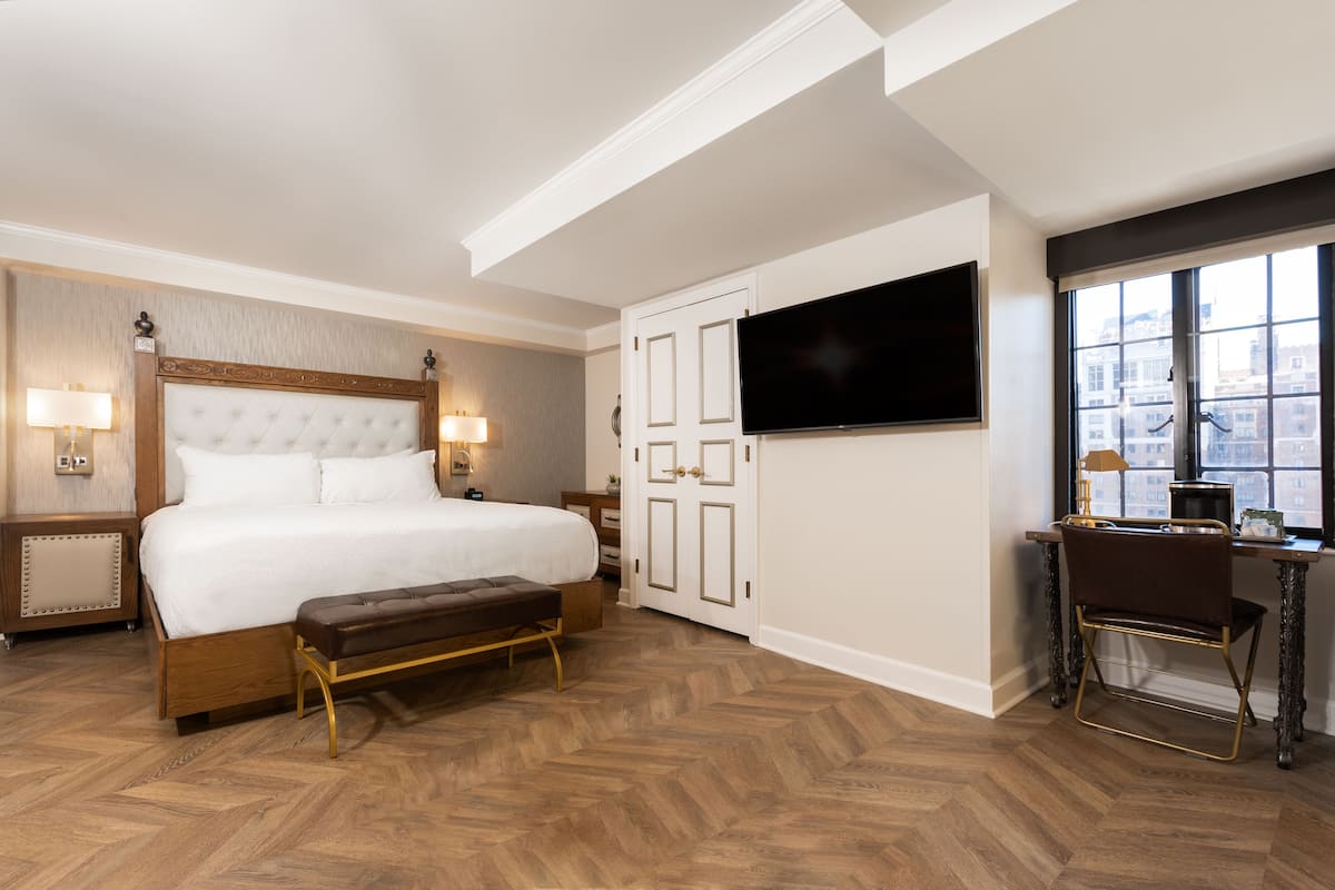 A hotel room with a large bed, a desk with a chair, a TV, and a window with a city view.