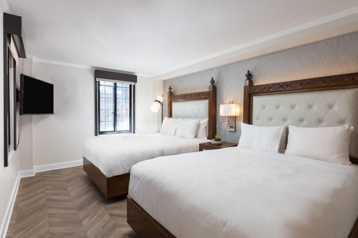 A hotel room with two beds, a TV, a window, and a wall-mounted lamp.