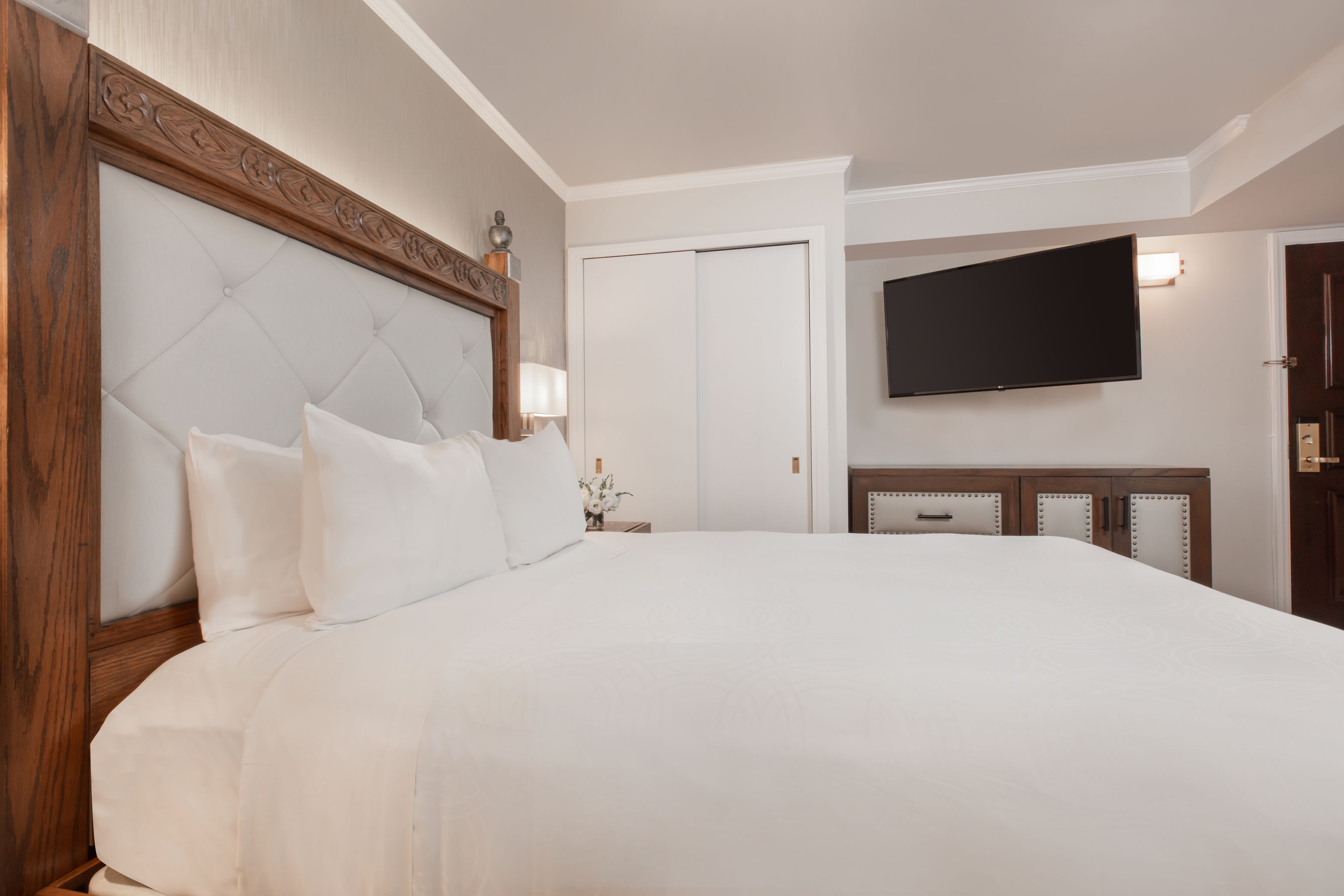 A large bed with a wooden headboard, a flat-screen TV mounted on the wall, and a closed door in the background.