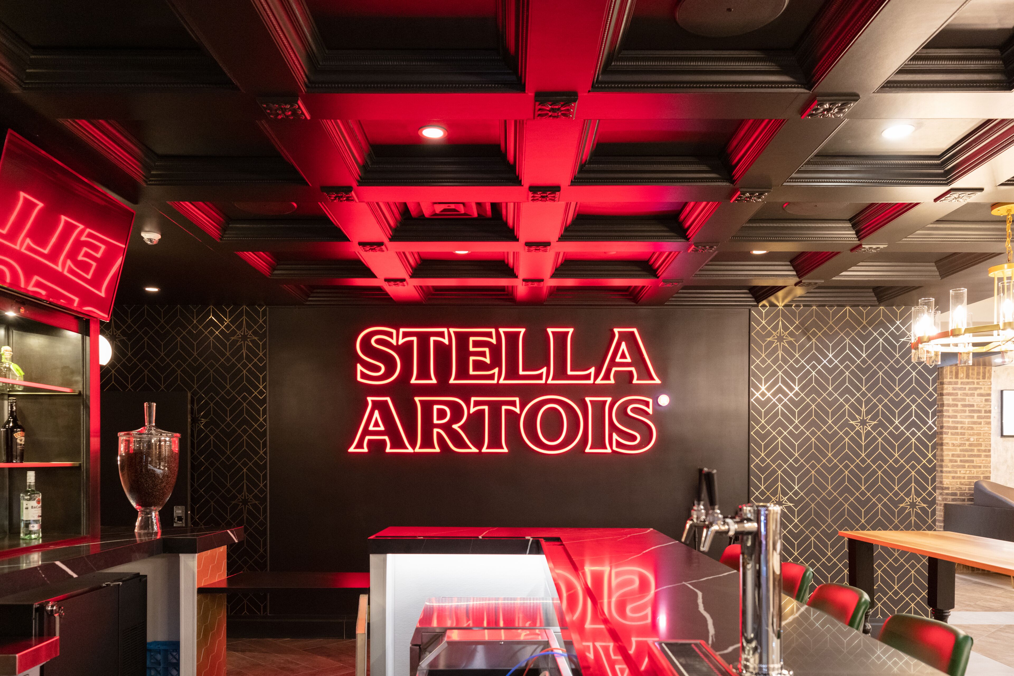 L’intérieur d’un restaurant, avec un panneau néon indiquant « STELLA ARTOIS », un bar où sont exposées des bouteilles, et un espace aménagé avec des tables et des chaises.