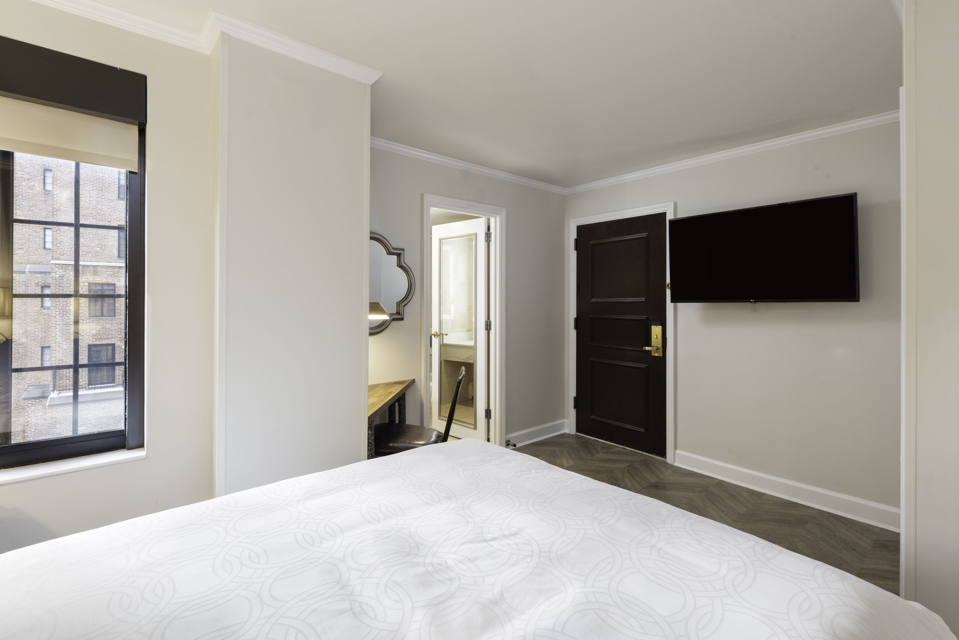 A hotel room with a large bed, a flat-screen TV mounted on the wall, and a view of the street through a window.