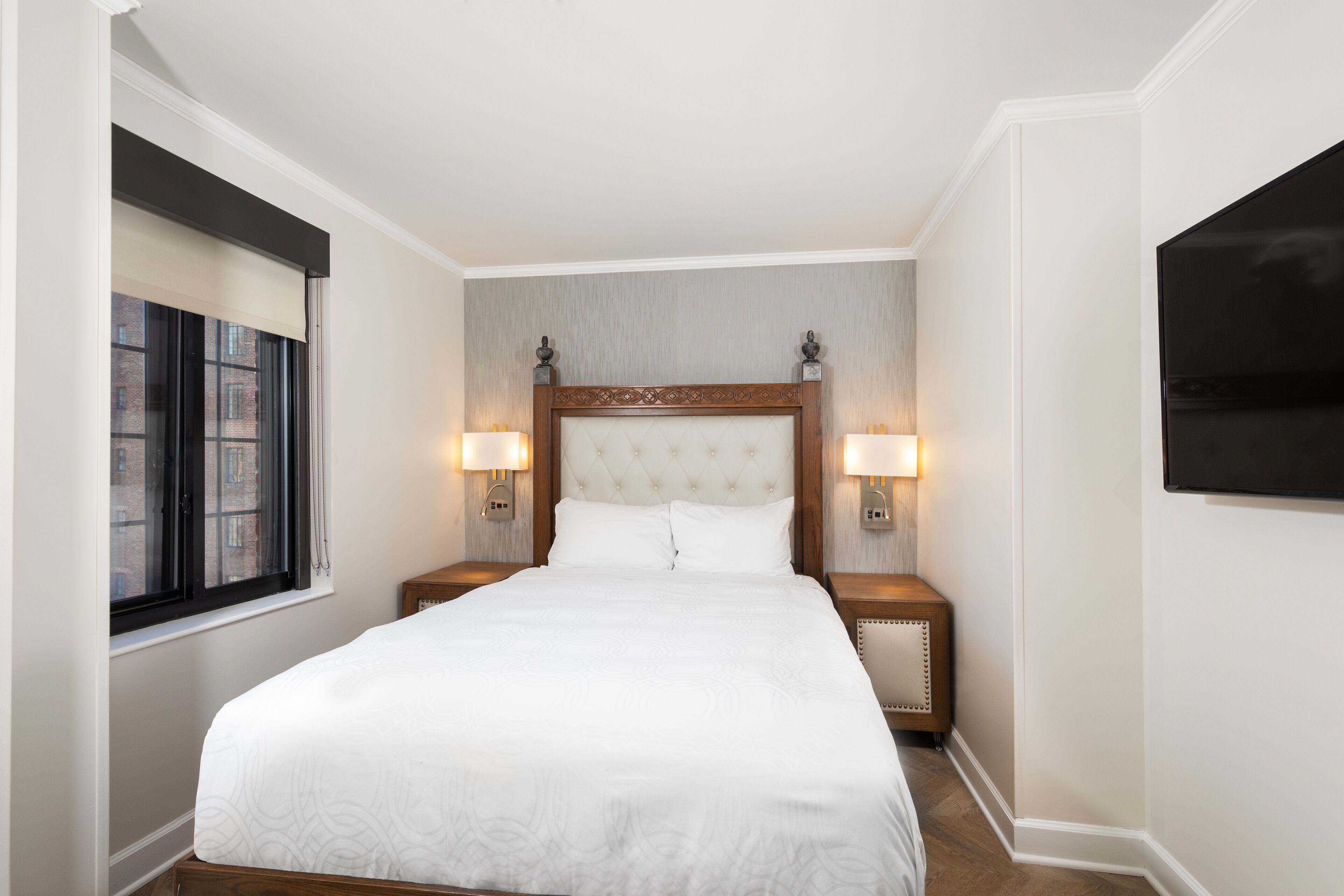 A neatly made bed with a tufted headboard, two bedside lamps, and a window with blinds.