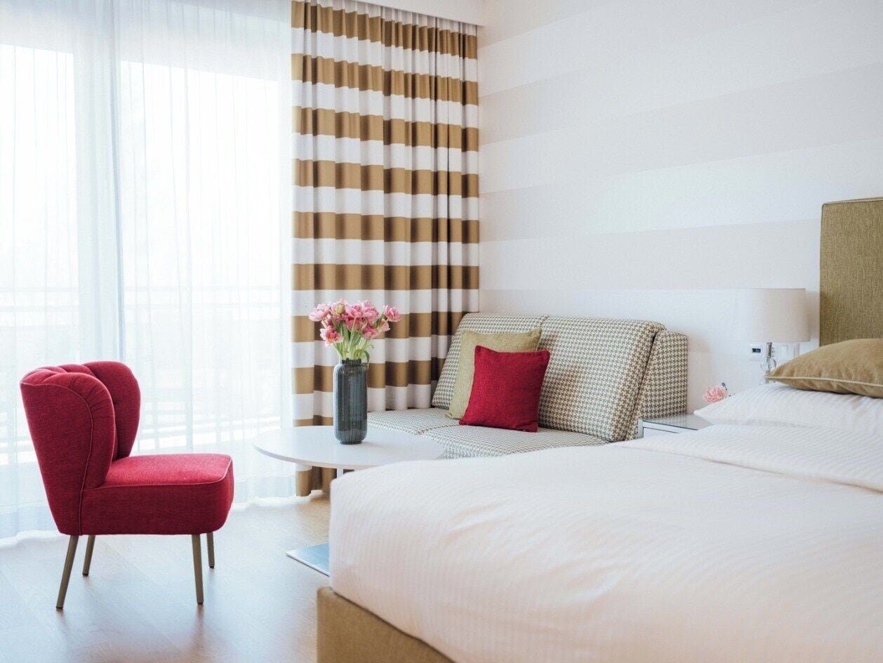 A hotel room with a large bed, a red armchair, a small round table, and a vase of flowers.