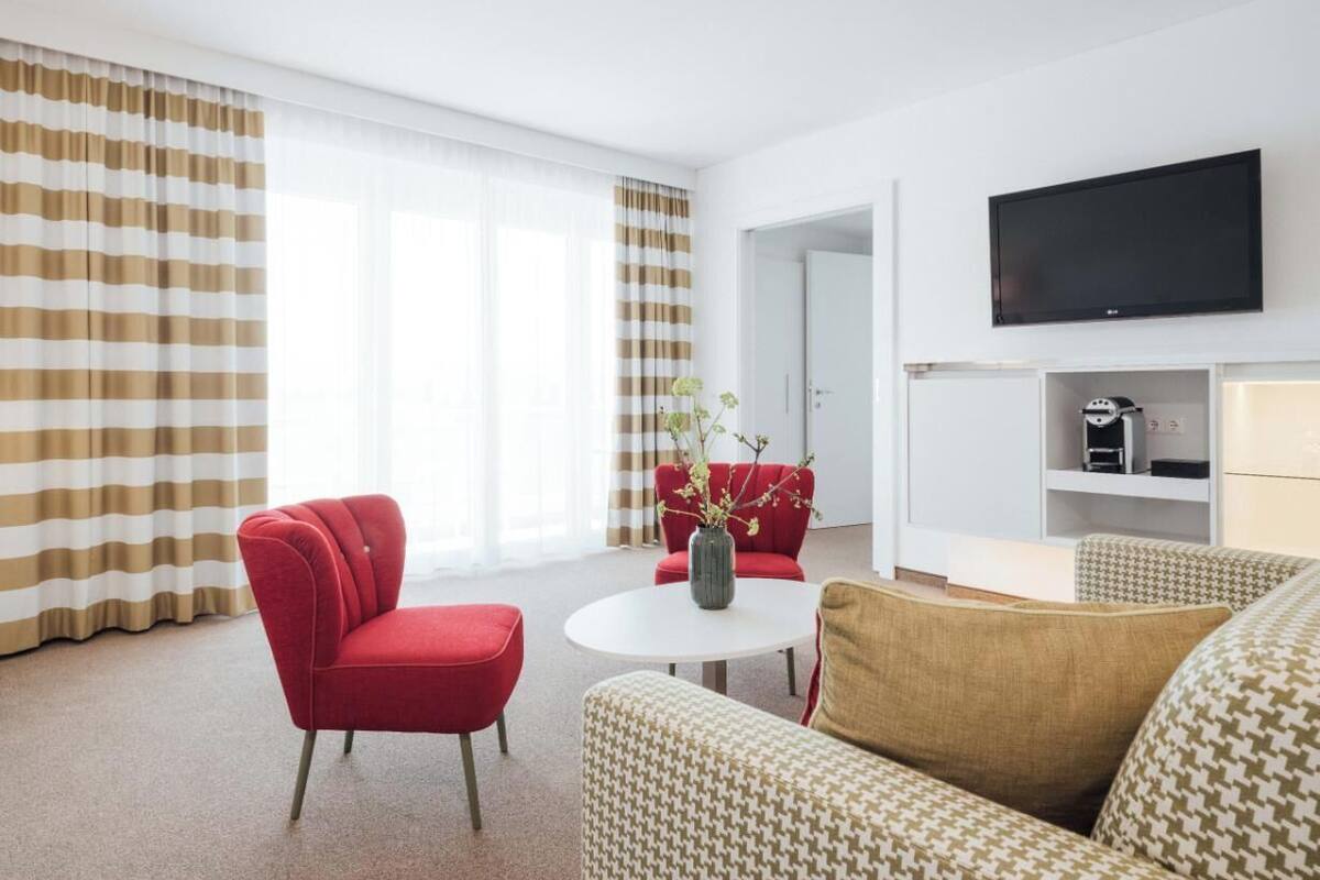 A hotel room with a red armchair, a patterned sofa, a white round table with a vase, and a TV mounted on the wall.