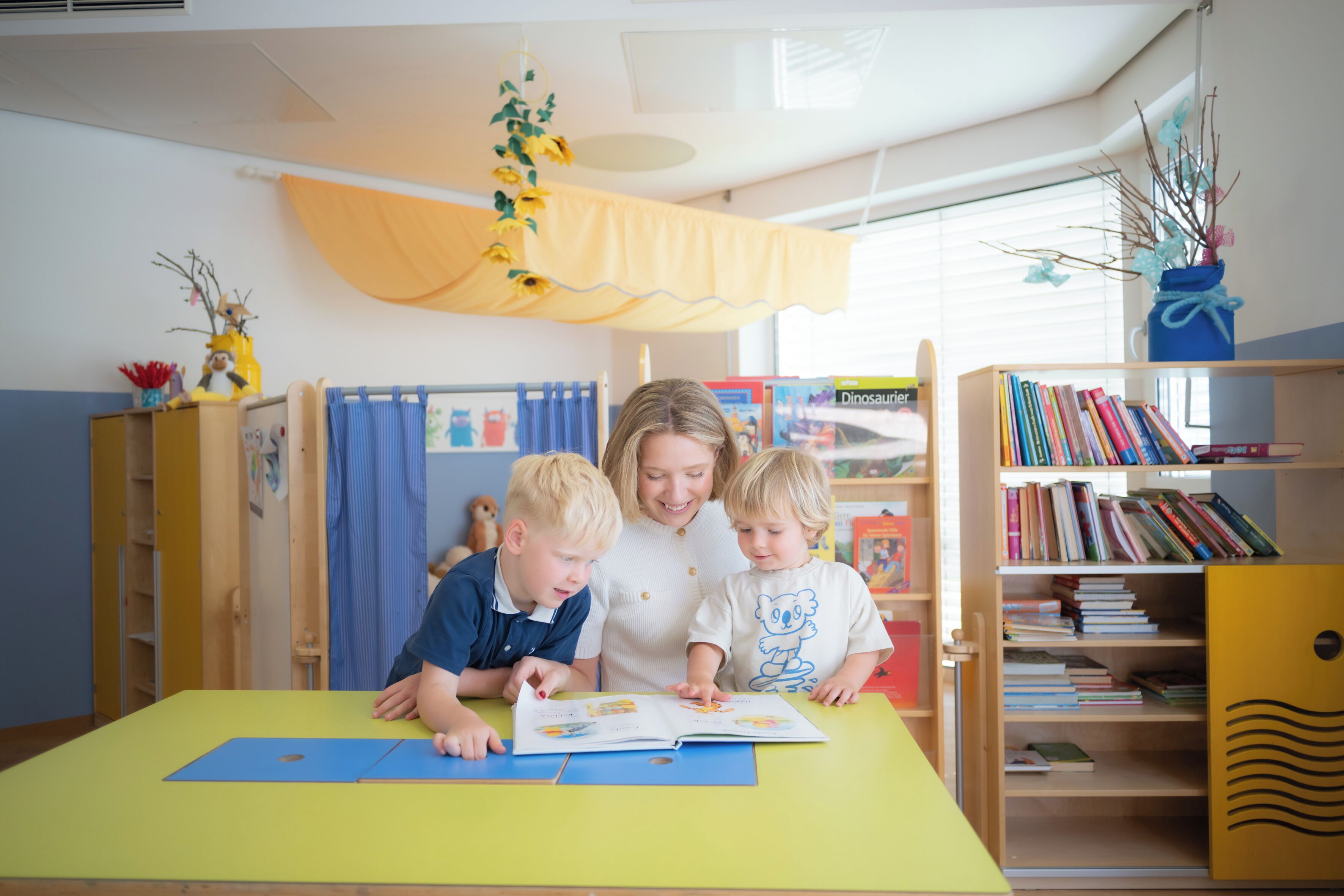 A room with a yellow table, a bookshelf, and a window with blinds.