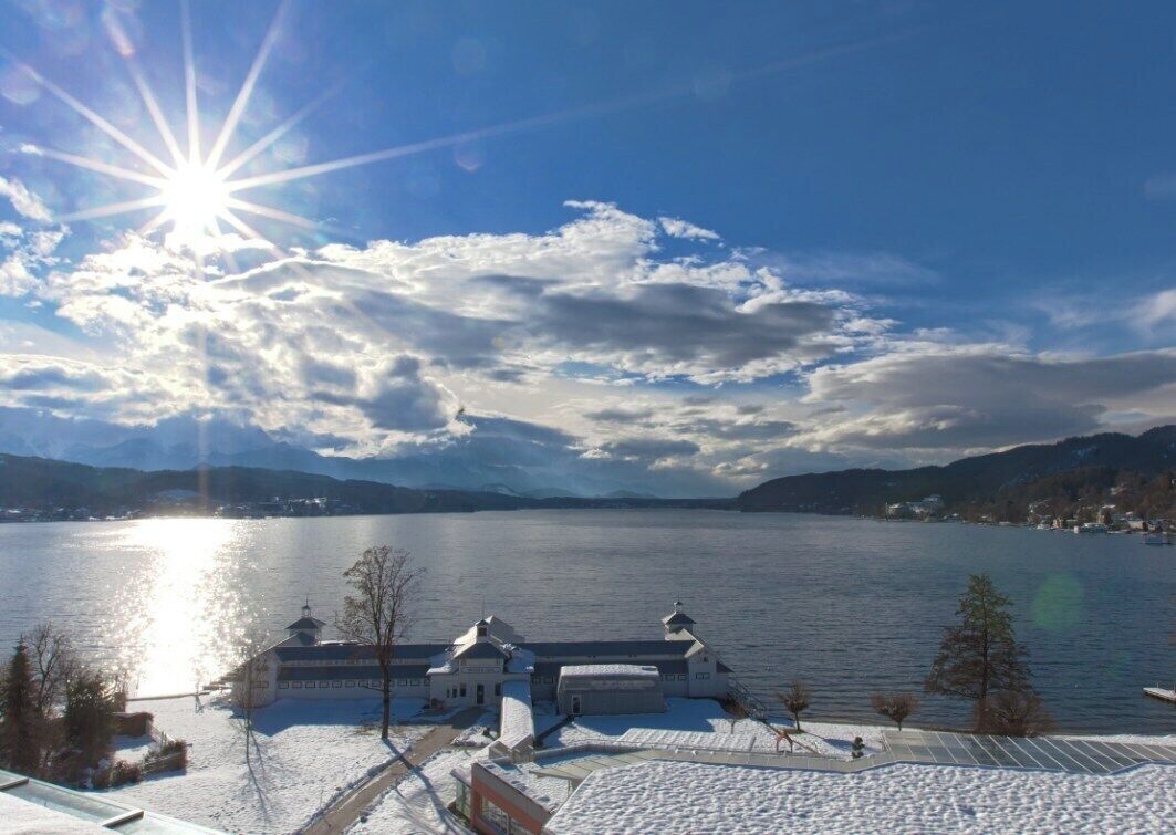 A lakeside view with a building featuring a large glass facade, a snow-covered ground, and mountains in the background.