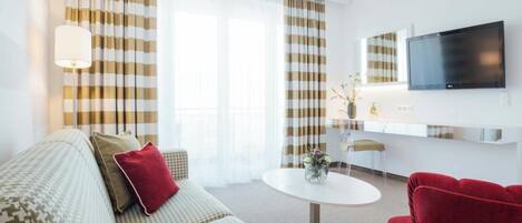 A modern hotel room with a sofa, a red armchair, a dining table, and a TV mounted on the wall.