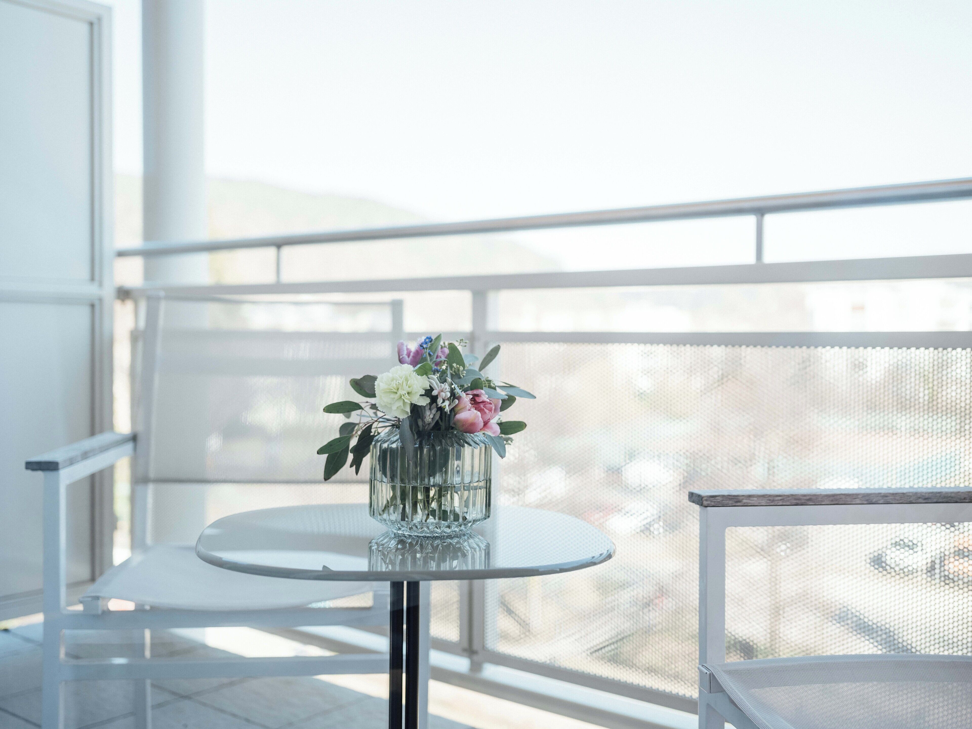 Un balcon avec une table ronde et des chaises, un vase en verre rempli de fleurs et une vue à travers un moustiquaire.
