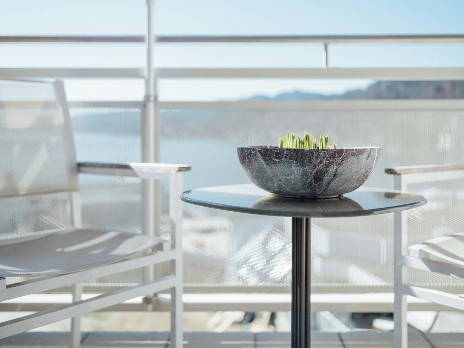 Un balcon moderne agrémenté d’un récipient en marbre contenant des plantes vertes, de chaises blanches et offrant une vue sur la mer.