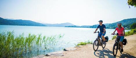 Two cyclists on a dirt path by a lake with mountains in the background.