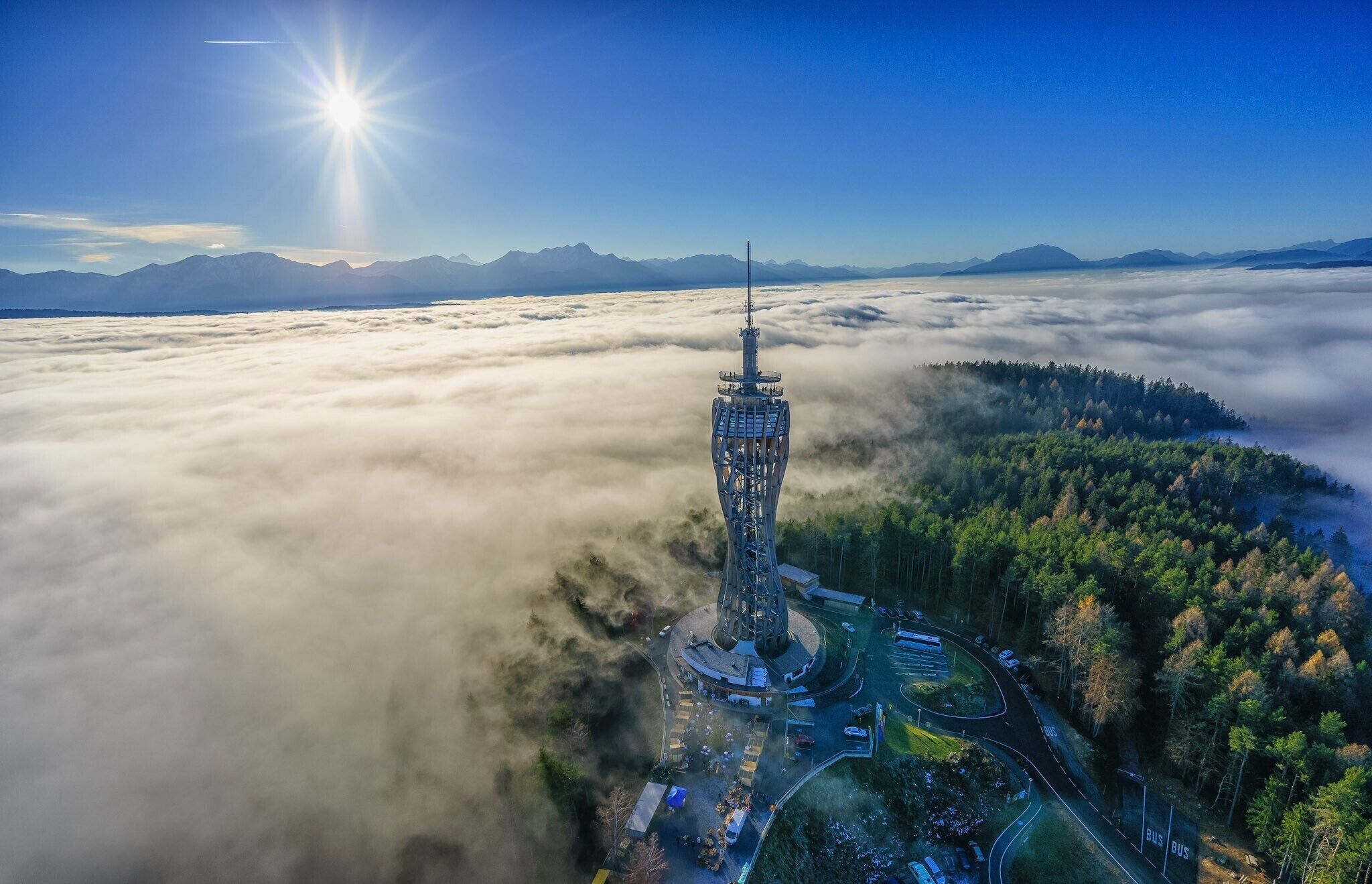 A tower surrounded by a forest and clouds, with a clear sky and mountains in the background.