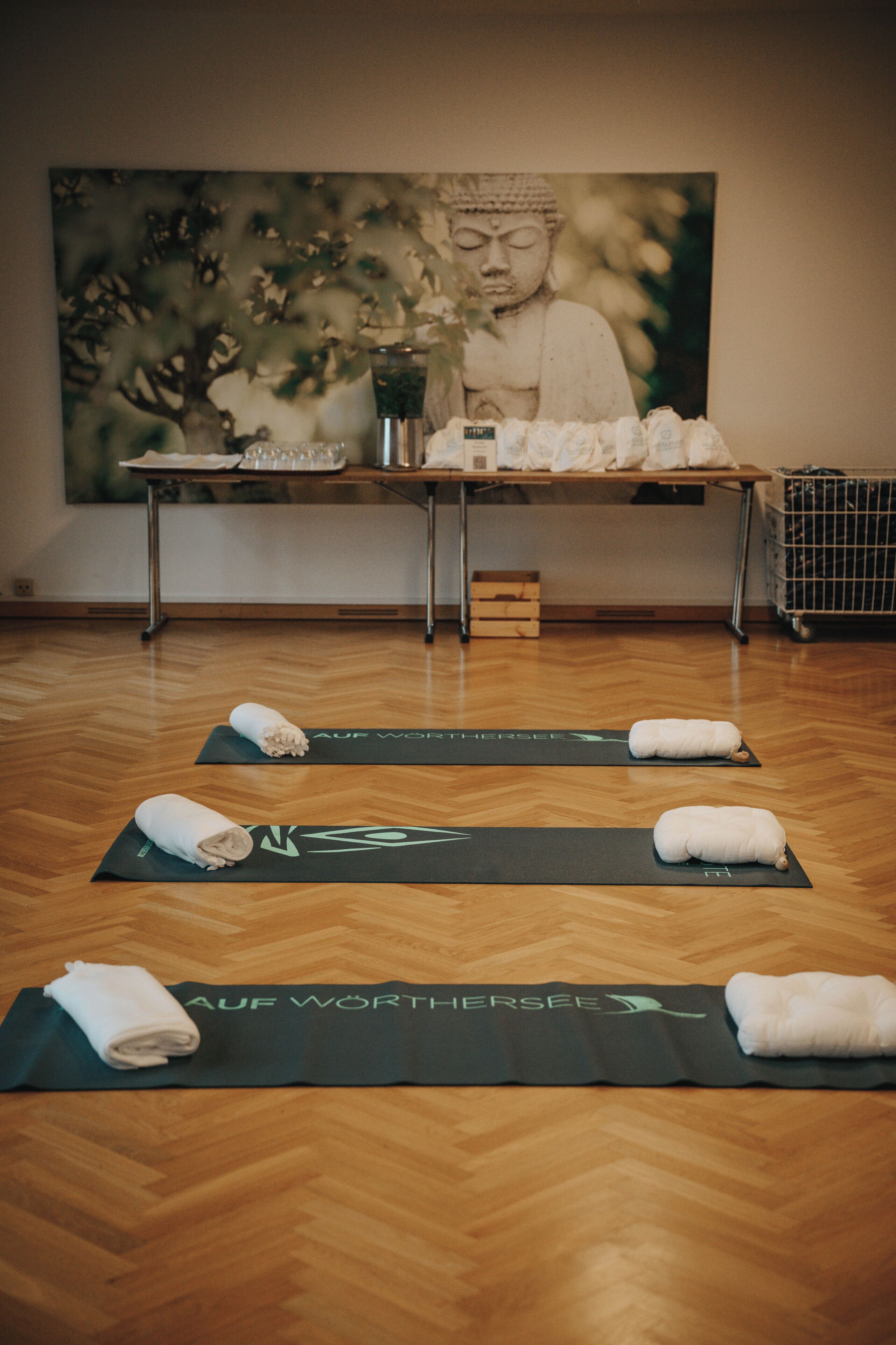 A room with wooden flooring, yoga mats, towels, a table with a Buddha statue, and a painting of a Buddha.