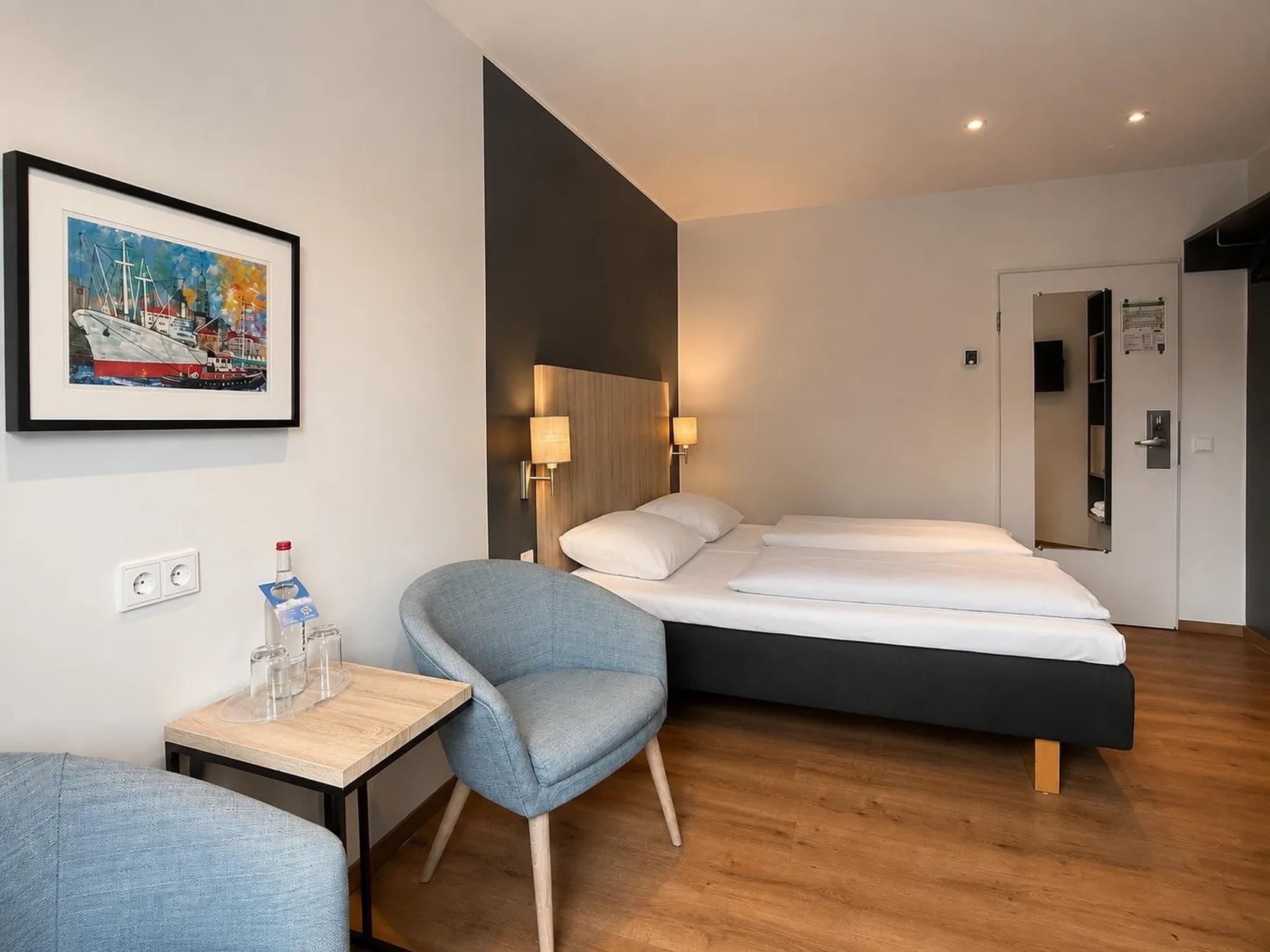 Ein Hotelzimmer mit einem Bett, einem Sessel, einem kleinen Tisch mit Gläsern und einer Flasche sowie einem Gemälde an der Wand.