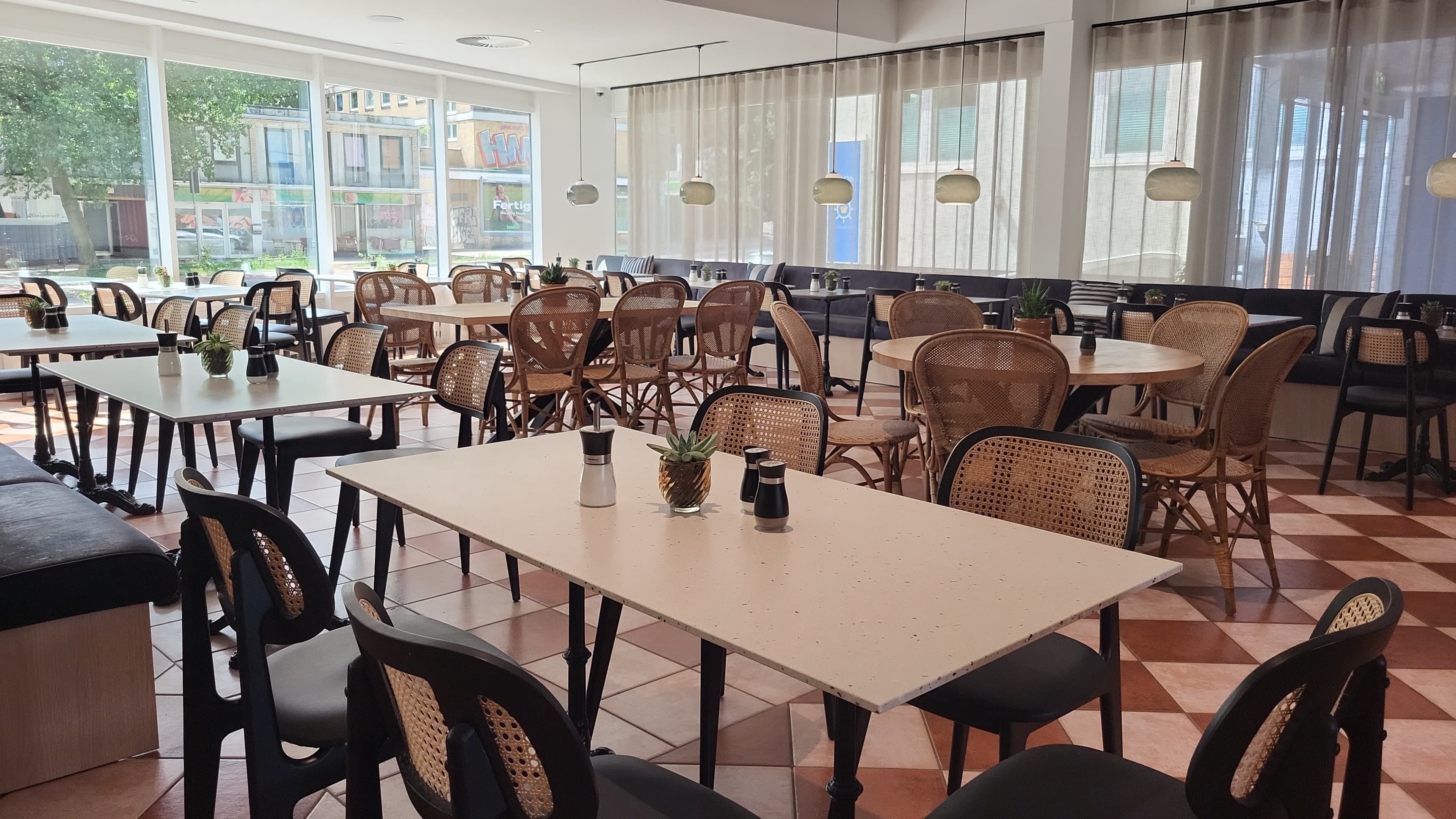 A spacious indoor dining area with multiple tables and chairs, large windows with curtains, and hanging pendant lights.