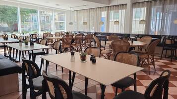 A spacious indoor dining area with multiple tables and chairs, large windows with curtains, and hanging pendant lights.