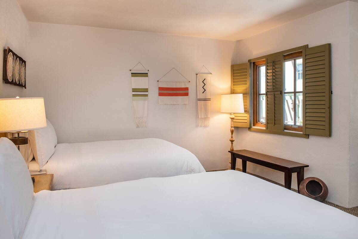 A hotel room with two beds, a wooden bench, a wall-mounted lamp, and a window with shutters.