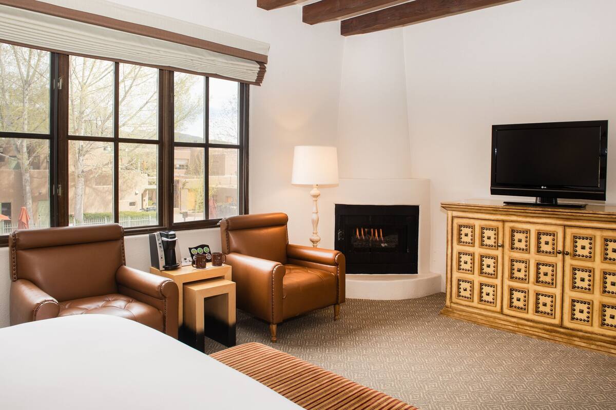 A hotel room with a large window, a fireplace, a television, and leather armchairs.