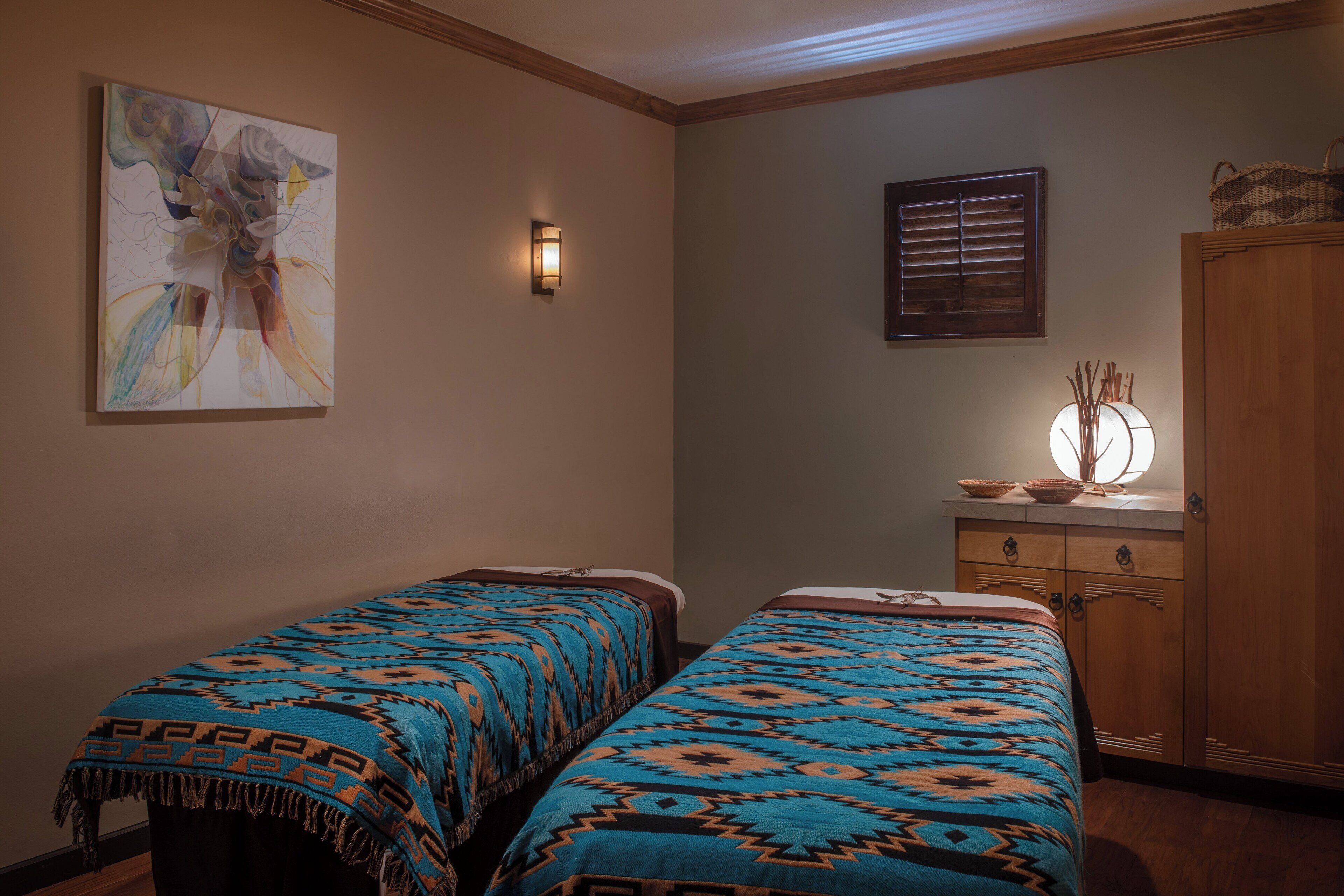 A room with two beds, a wall-mounted light, a window with blinds, and a wooden cabinet.