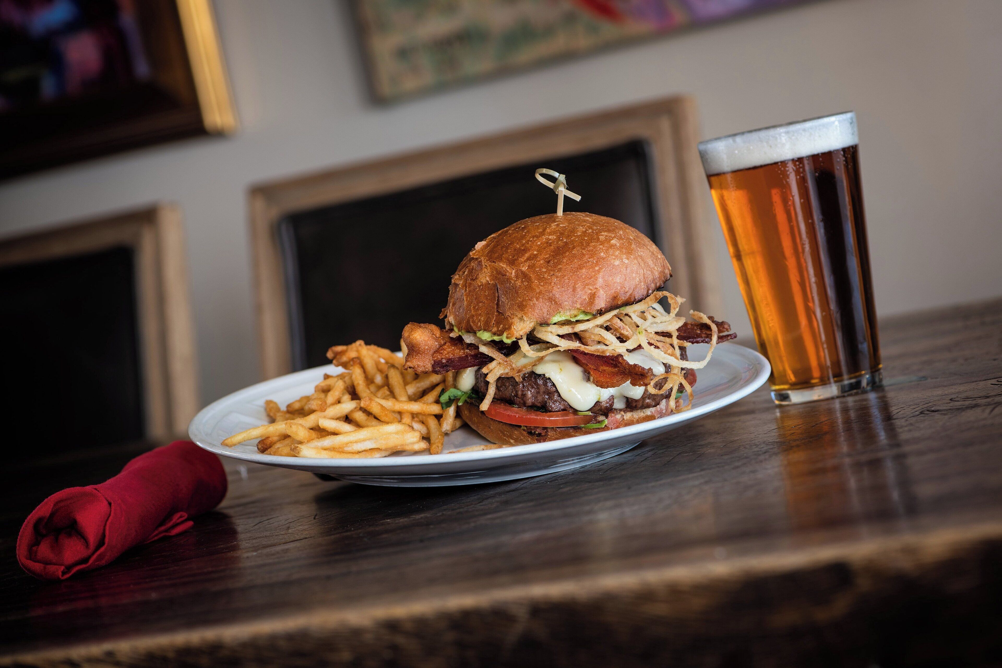 Un plato con una hamburguesa y papas fritas, un vaso de cerveza y una servilleta roja sobre una mesa de madera.