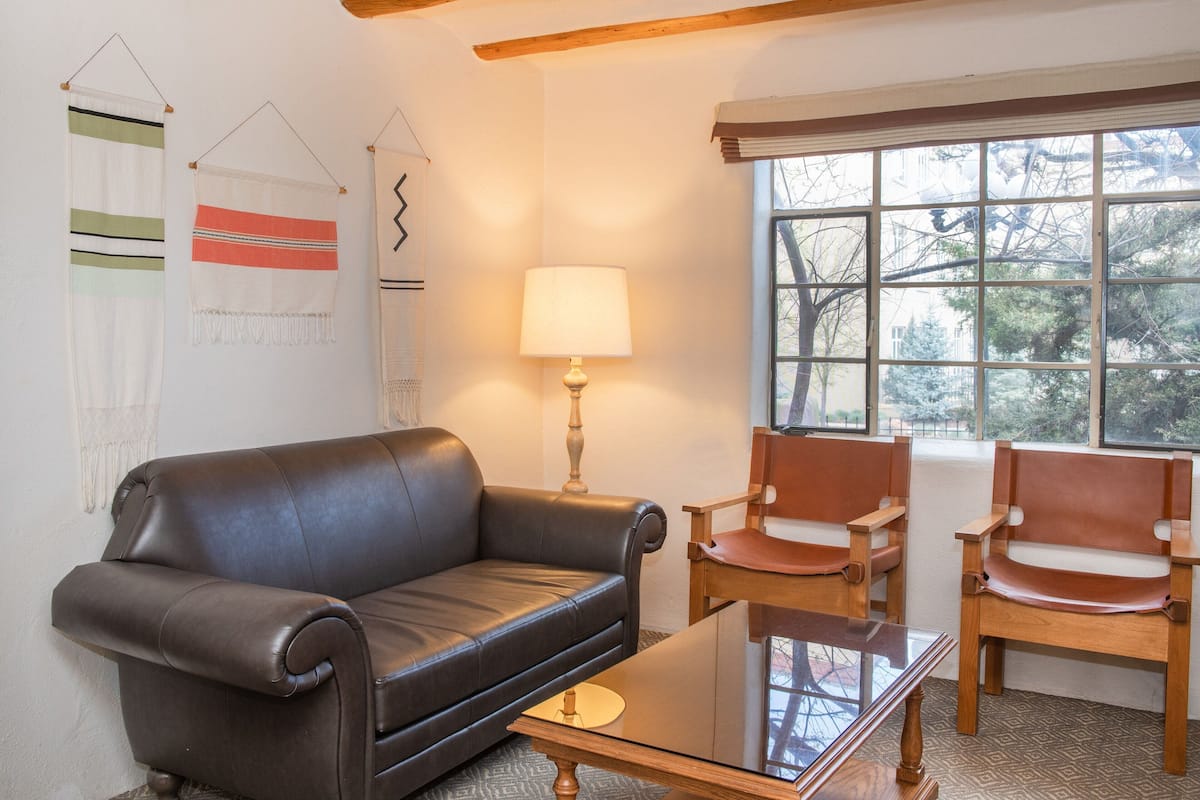 A living room with a leather sofa, two wooden chairs, a glass coffee table, a lamp, and a window with a view of trees.