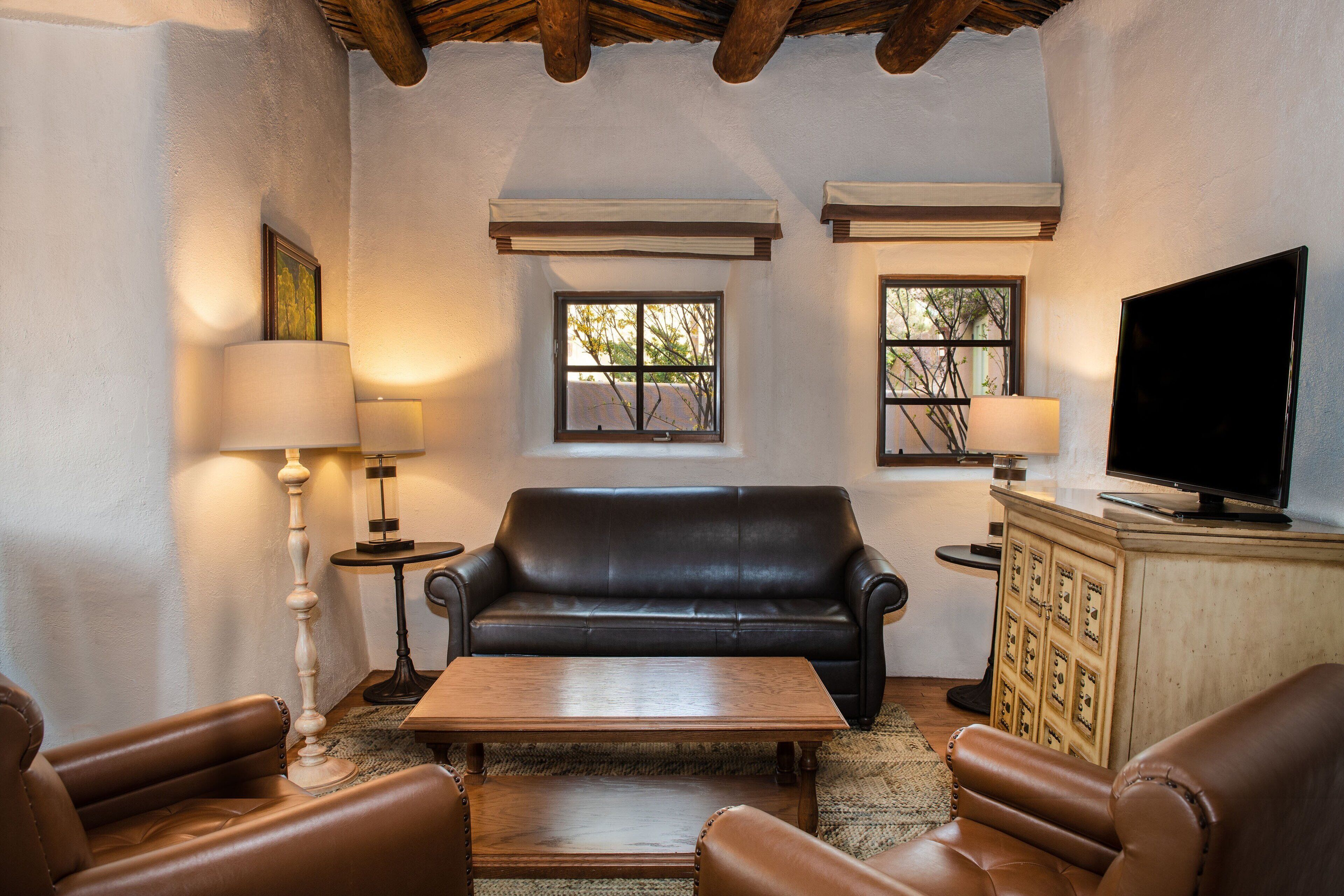 A living room with a black leather sofa, a wooden coffee table, and leather armchairs. There are two windows with blinds, a television on a cabinet, and a lamp on a side table.