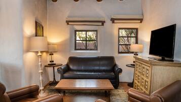 A living room with a black leather sofa, a wooden coffee table, and leather armchairs. There are two windows with blinds, a television on a cabinet, and a lamp on a side table.