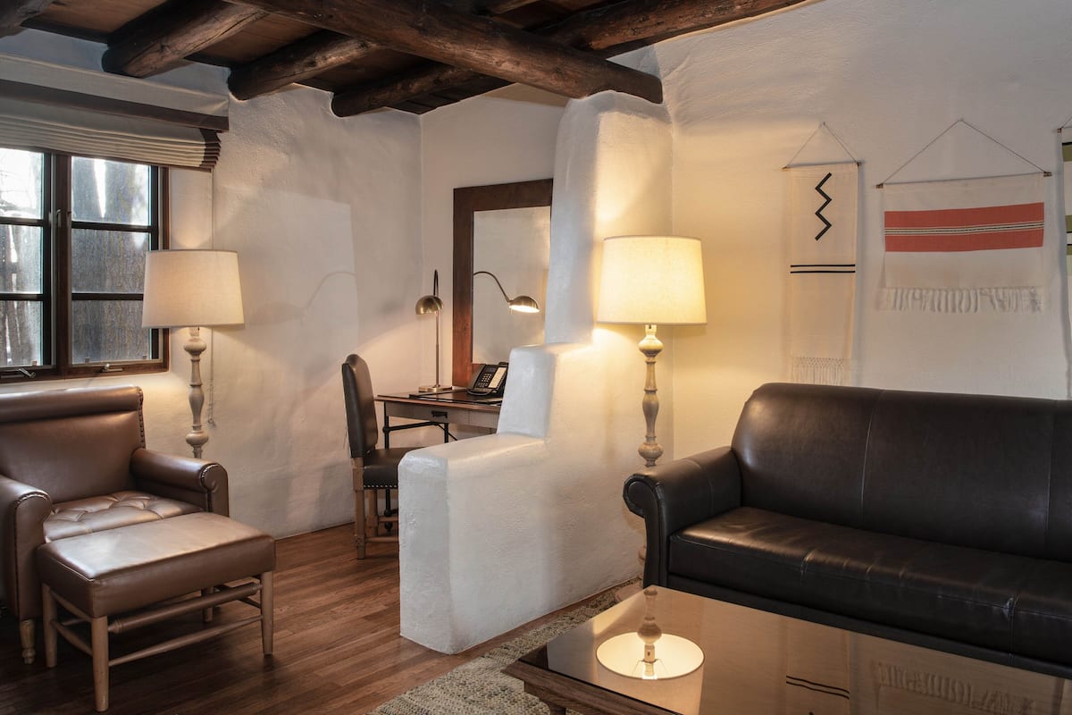 A cozy living room with a leather sofa, a cushioned armchair, and a glass coffee table. There is a desk with a chair and a lamp. The room has wooden beams on the ceiling and a window with blinds.