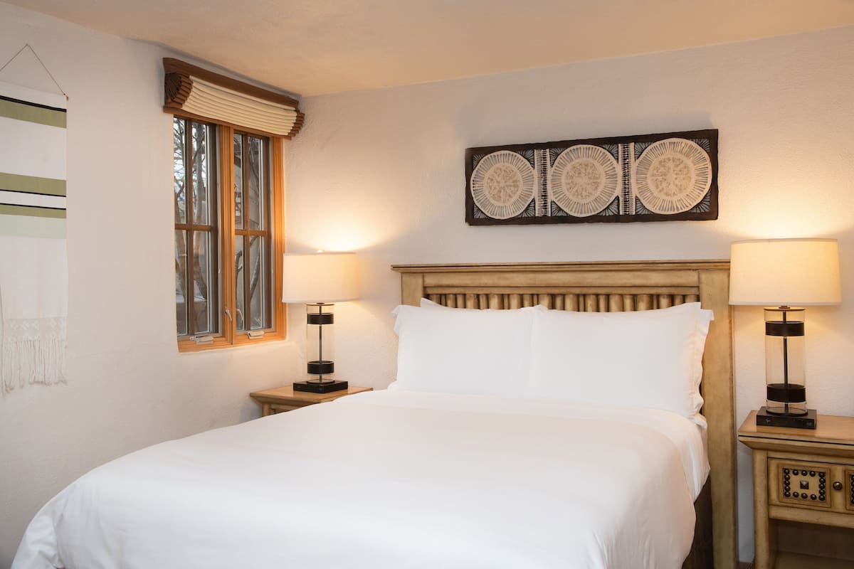 A neatly made bed with white linens, two bedside tables with lamps, a window with a wooden frame, and a wall-mounted decorative panel.