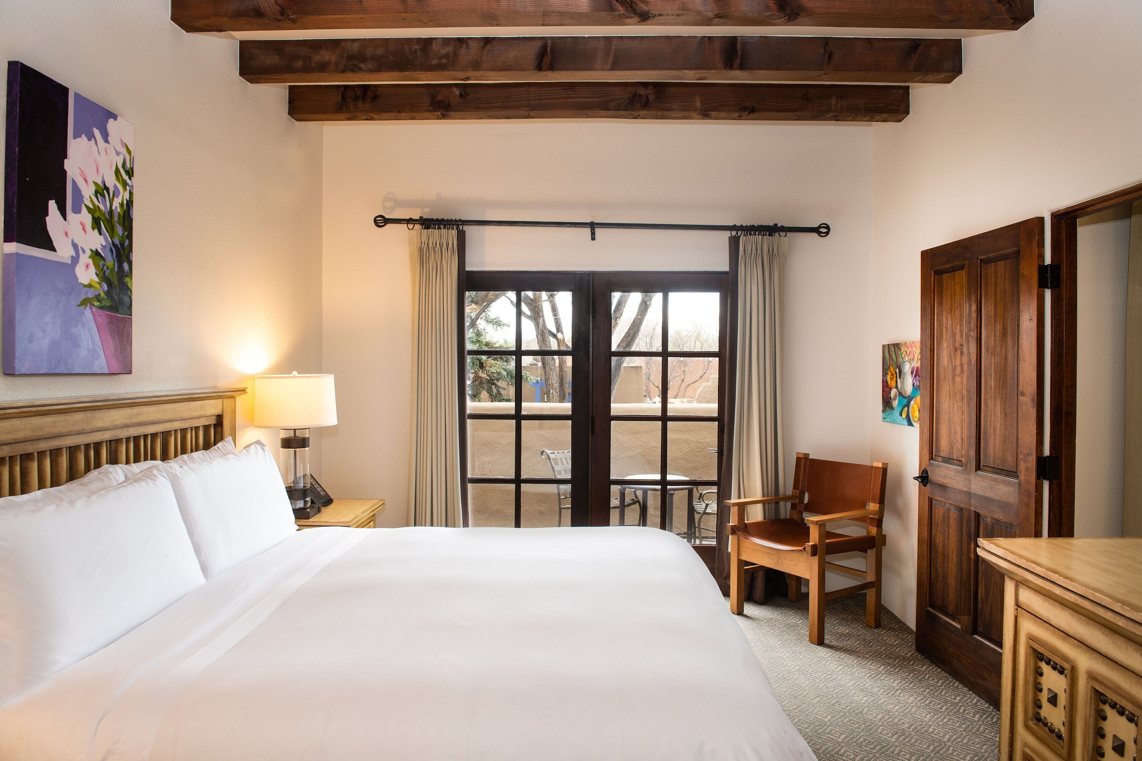 A hotel room with a large bed, a wooden headboard, a nightstand with a lamp, a chair, and a wooden wardrobe.