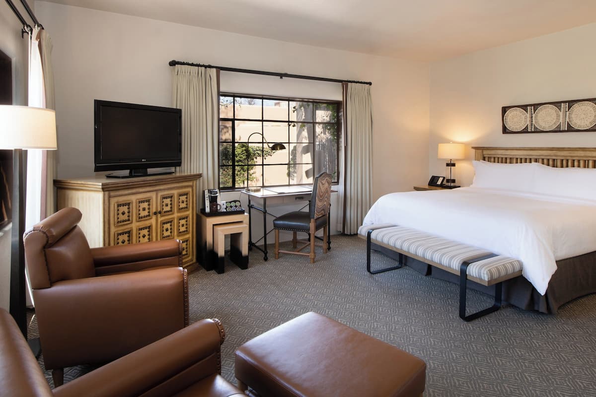 A hotel room with a large bed, a desk with a chair, a television, and a seating area with chairs and an ottoman.