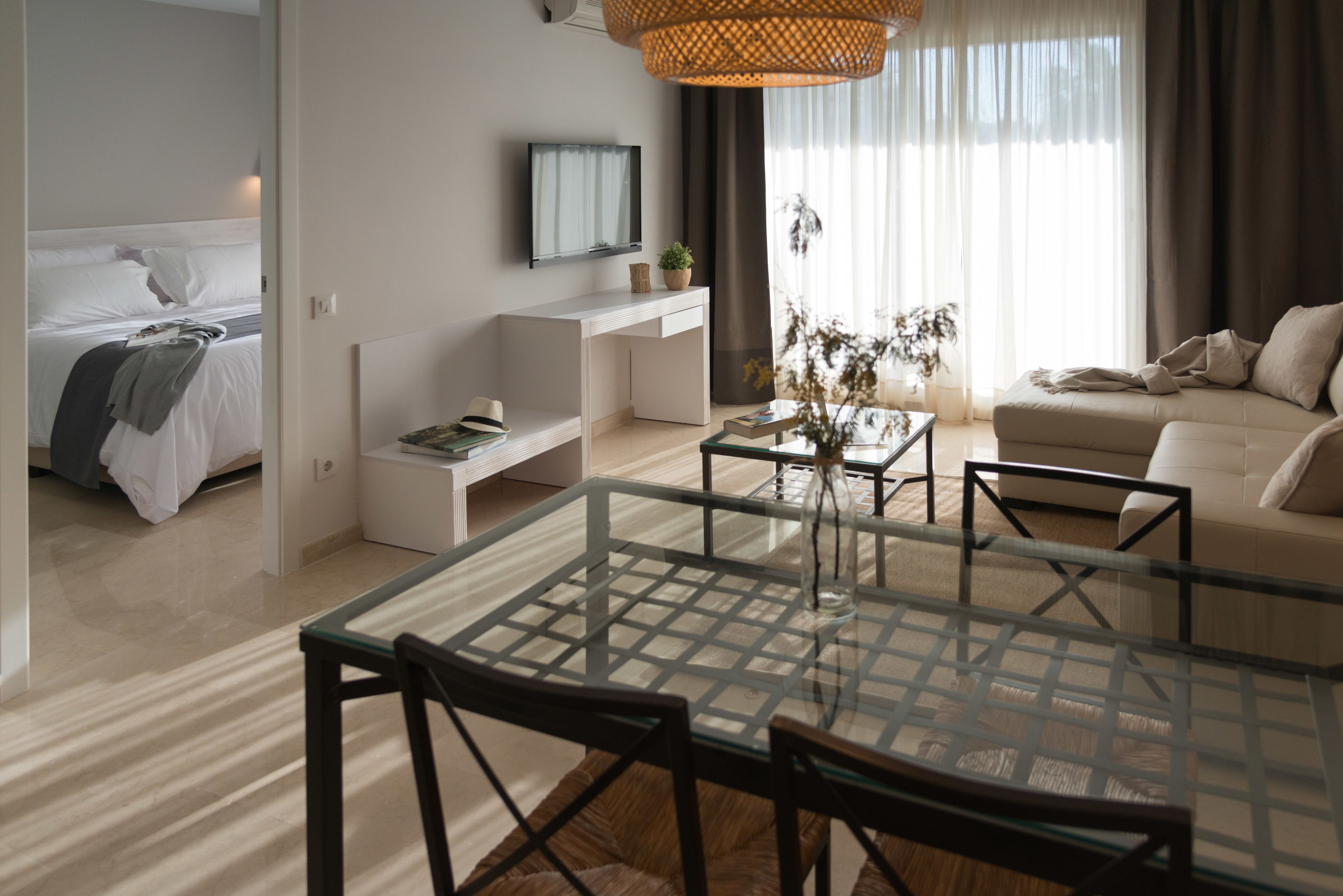 Una habitación de hotel moderna con una mesa de comedor de vidrio, un sofá, una cama, un televisor y un amplio ventanal con cortinas.