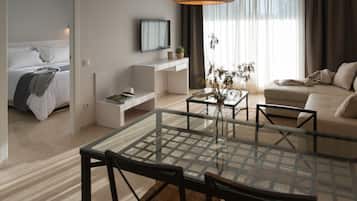 Una habitación de hotel moderna con una mesa de comedor de vidrio, un sofá, una cama, un televisor y un amplio ventanal con cortinas.