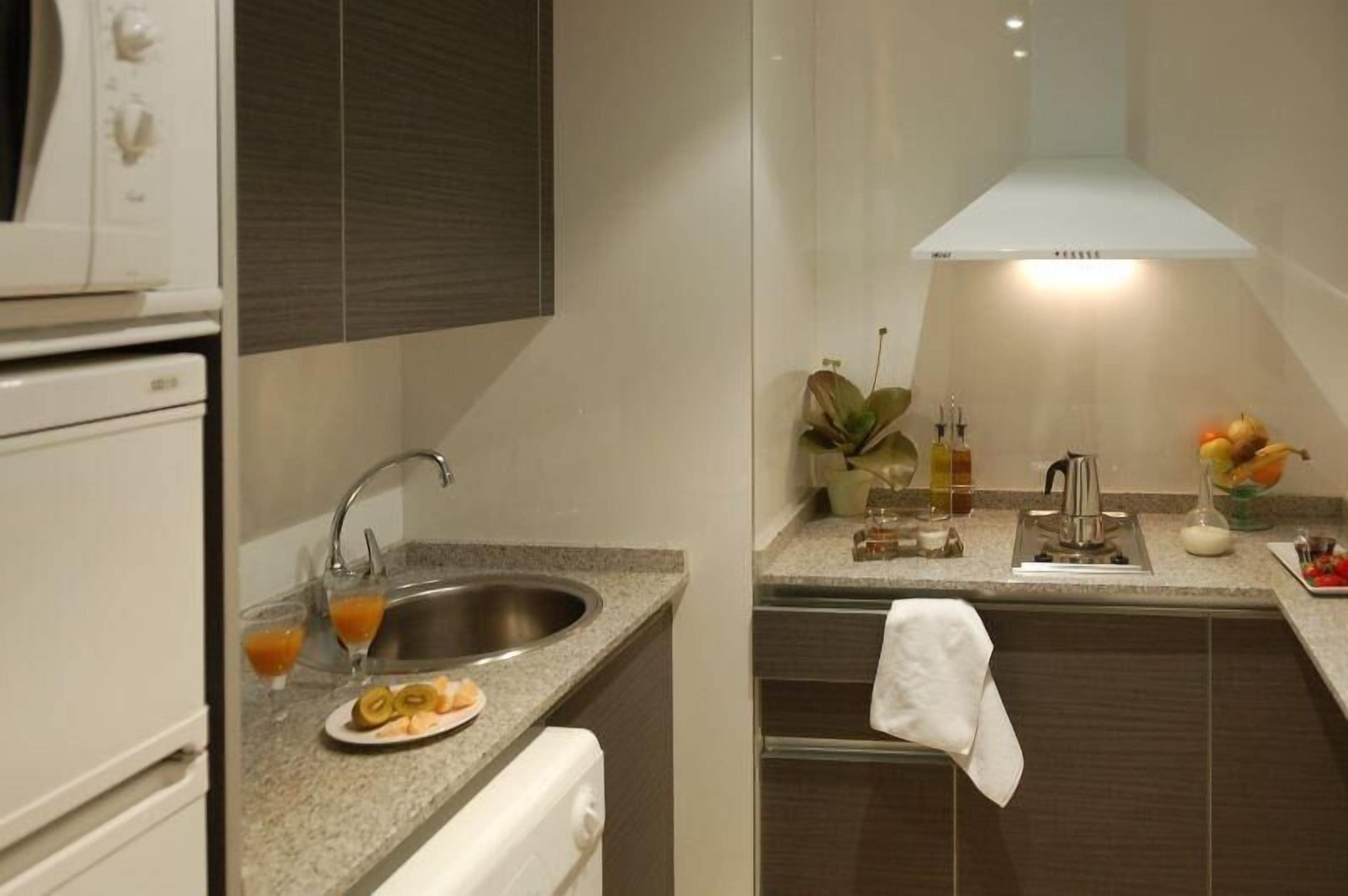 Una cocina moderna con un lavamanos de acero inoxidable, un horno empotrado y una campana extractora. Hay dos vasos de jugo de naranja en la encimera y un plato con fruta cortada.