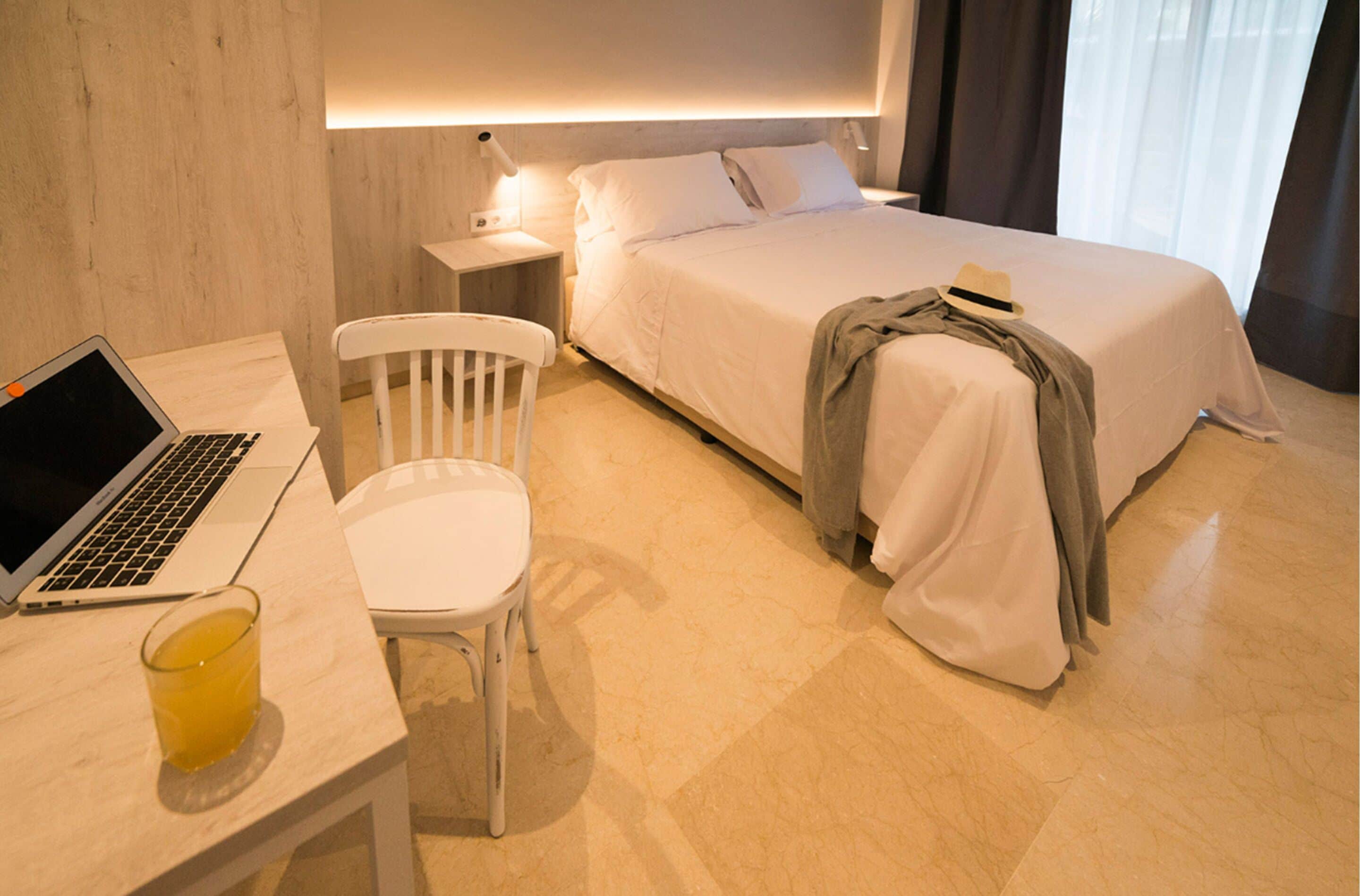 Quarto de hotel com cama, escrivaninha com laptop, cadeira e mesa de cabeceira com abajur.
