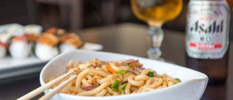 A bowl of noodles with chopsticks, a glass of beer, and a bottle of Asahi beer on a table.