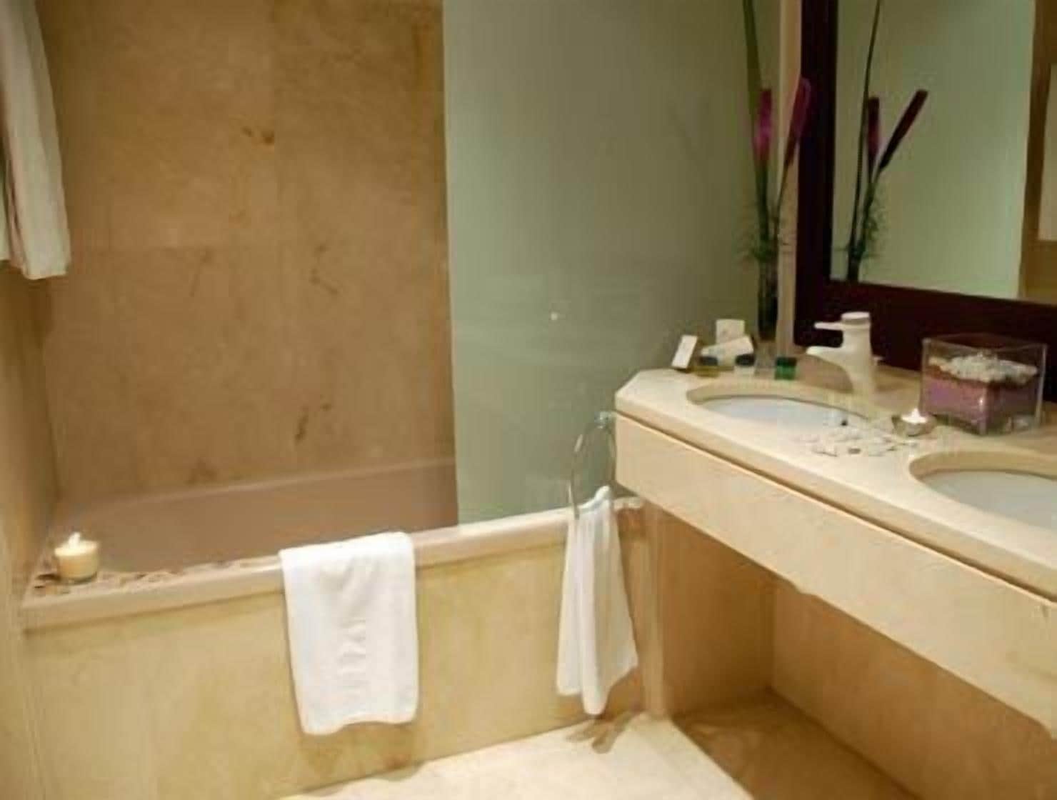 Un baño con bañera, lavamanos y espejo. Hay toallas colgadas en la pared y una vela sobre la bañera.