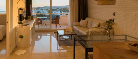 A modern living room with a sofa, coffee table, and a dining area. There is a balcony with a view of the city.