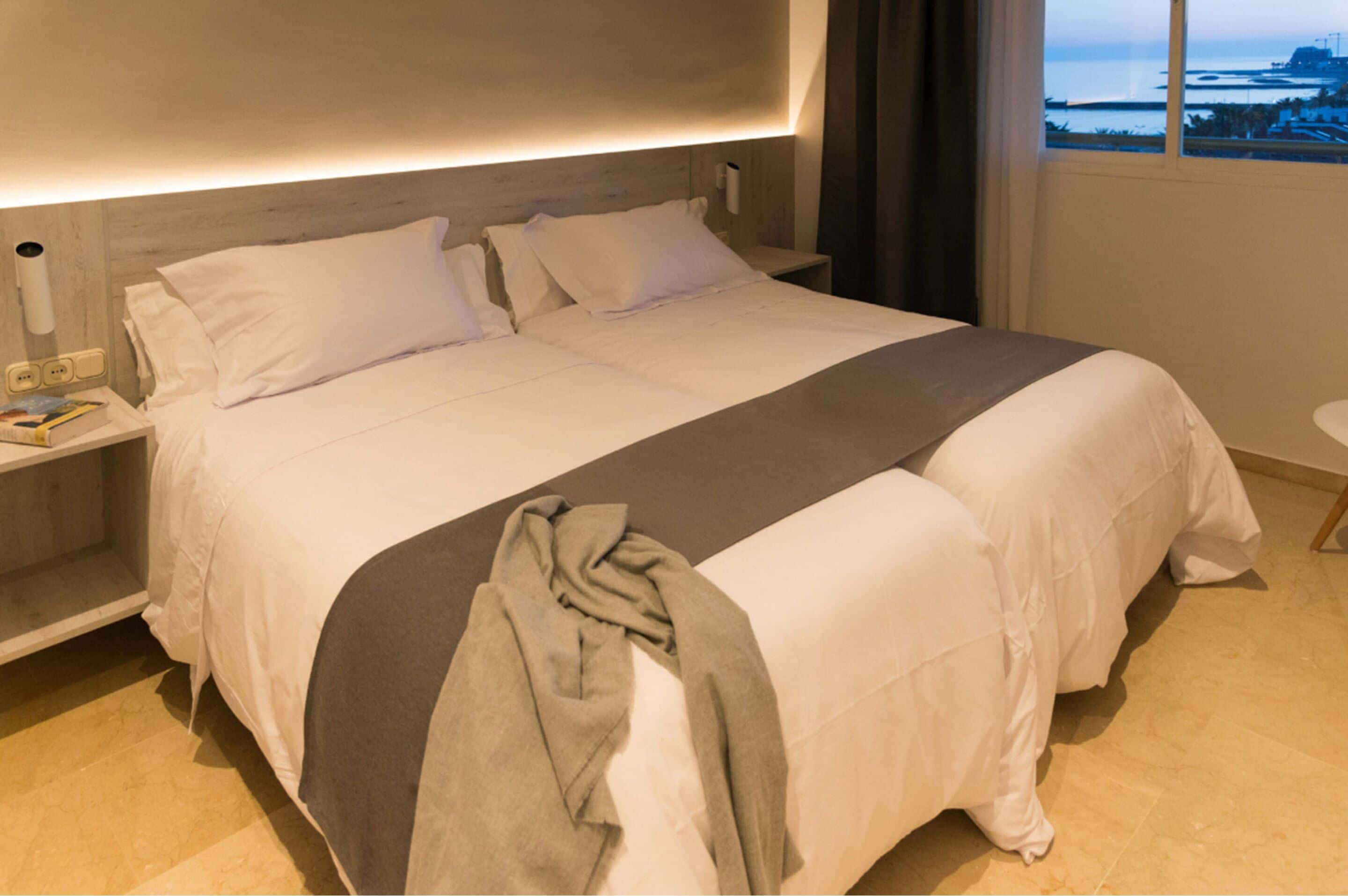 A large bed with white linens, a grey throw blanket, and a bedside table with a lamp.