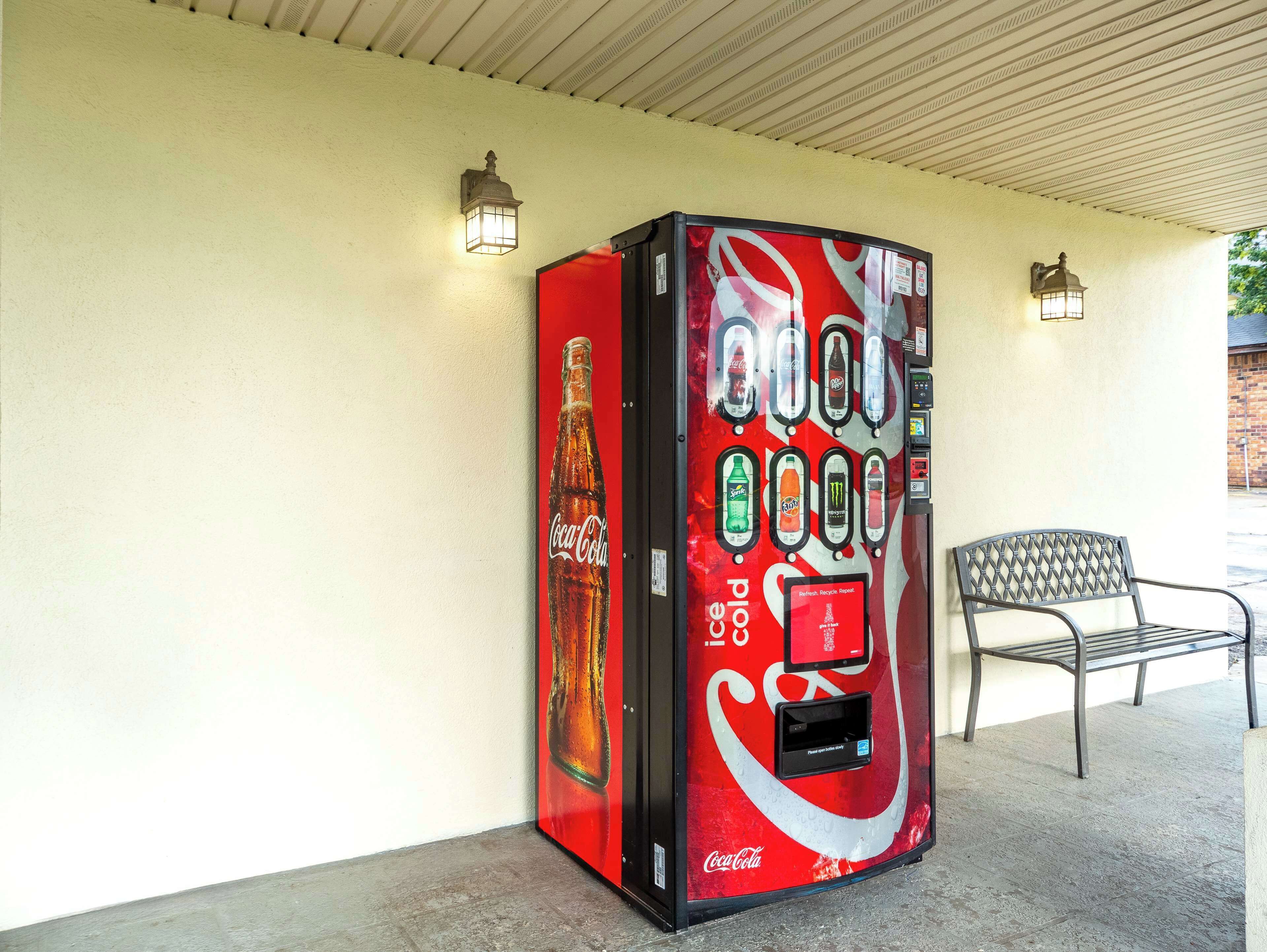 A Coca-Cola branded vending machine with a refreshment dispenser, located outdoors next to a bench.