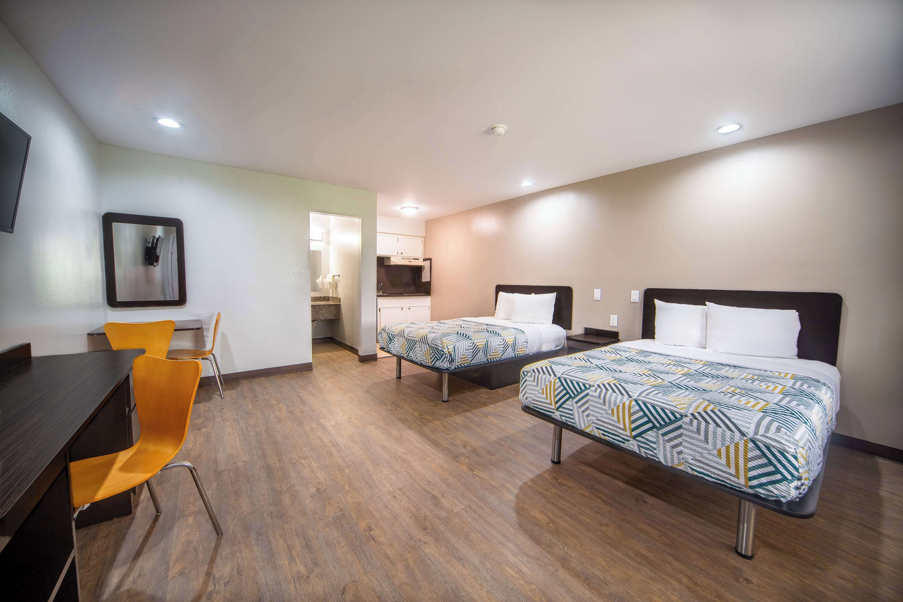A hotel room with two beds, a desk, and a chair. There is a kitchenette in the background.
