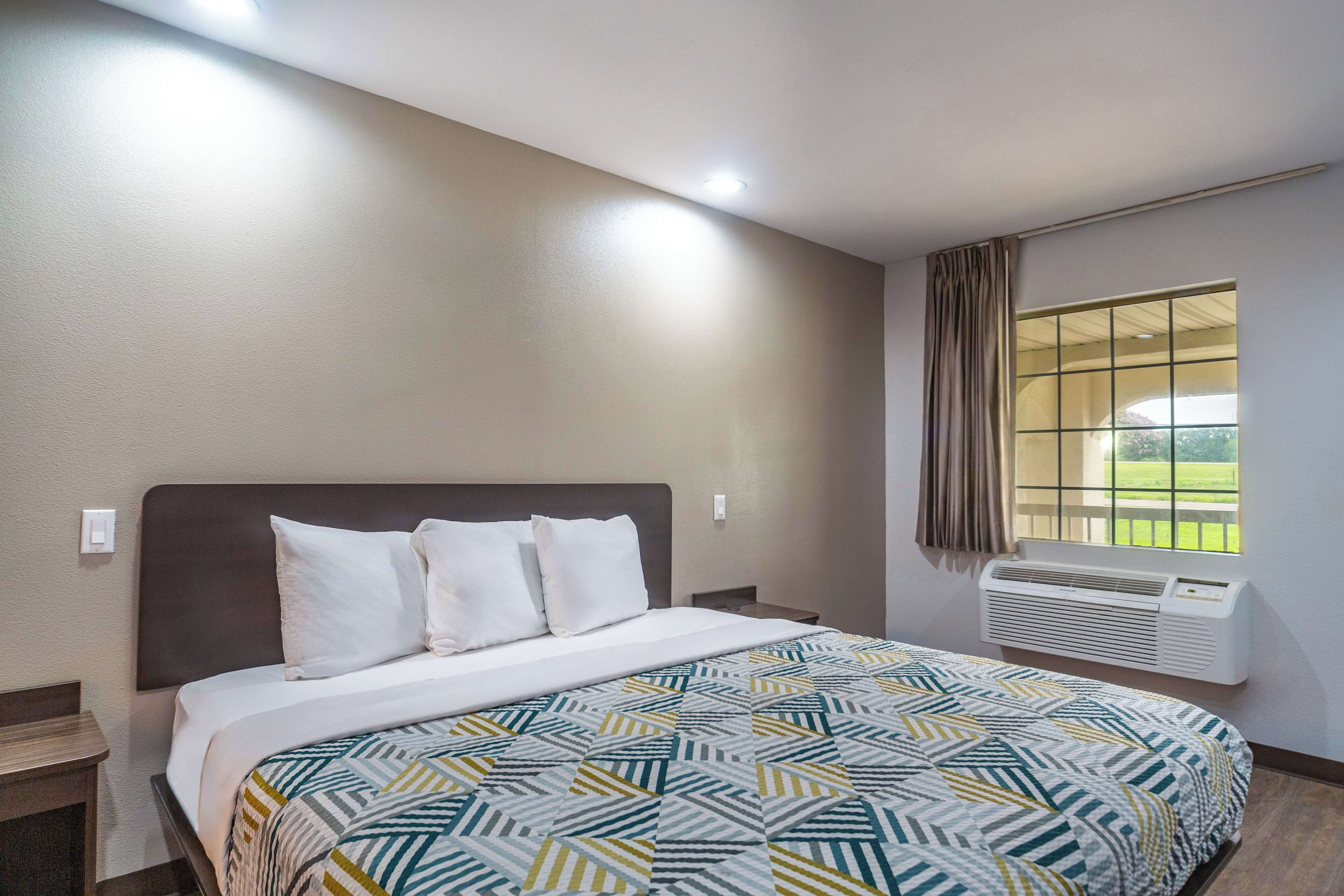 A hotel room with a bed, a window with curtains, an air conditioning unit, and a nightstand.