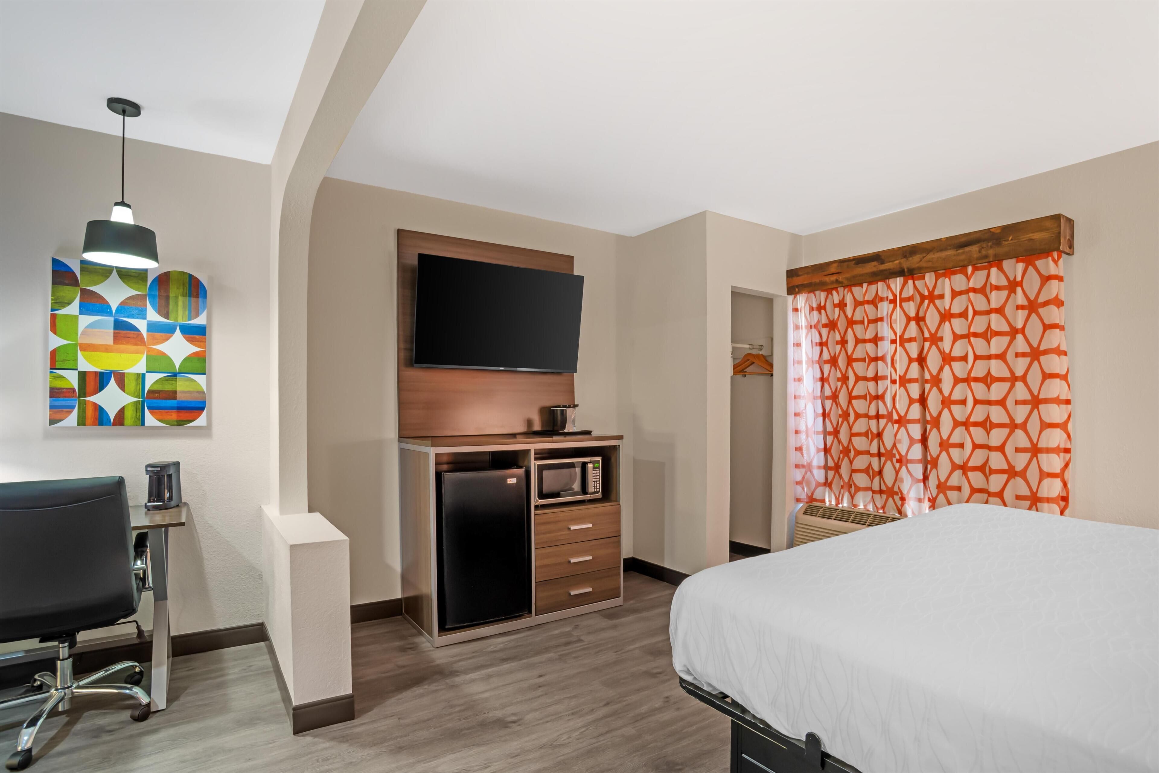 Une chambre d’hôtel équipée d’un lit, d’un bureau avec un ordinateur, d’un téléviseur à écran plat, d’un four à micro-ondes et d’un tableau abstrait aux couleurs vives.