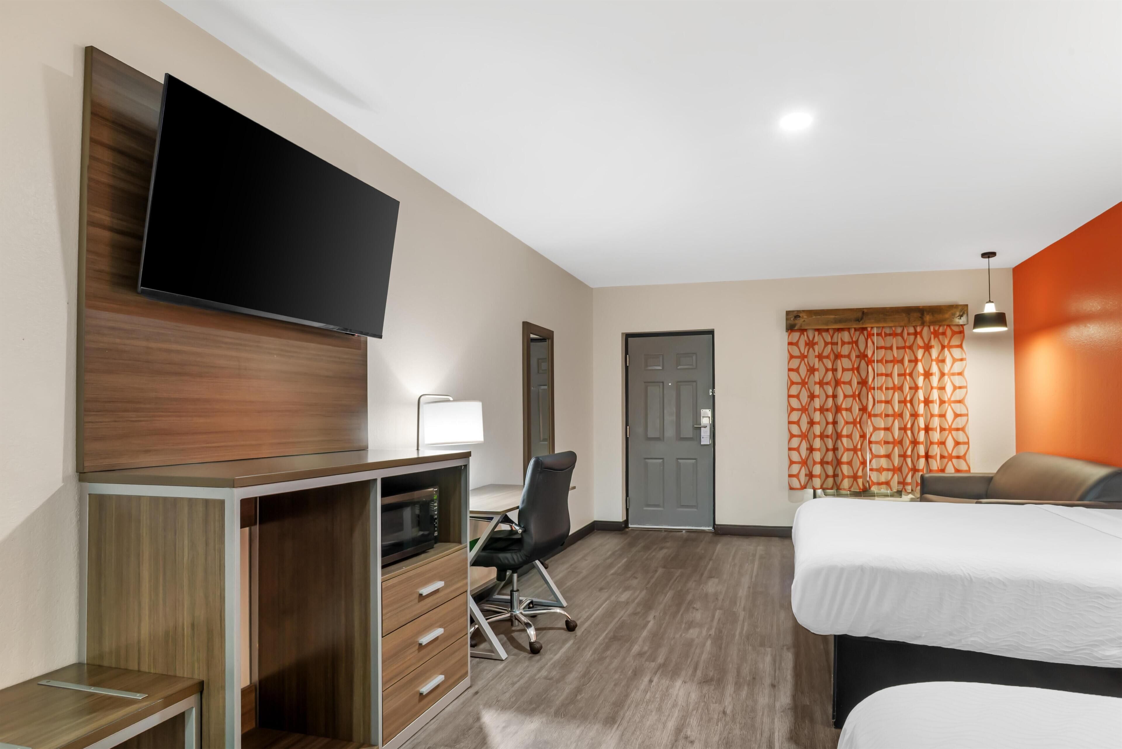 A hotel room with a flat-screen TV, a desk with a lamp, a chair, a bed, and a door.