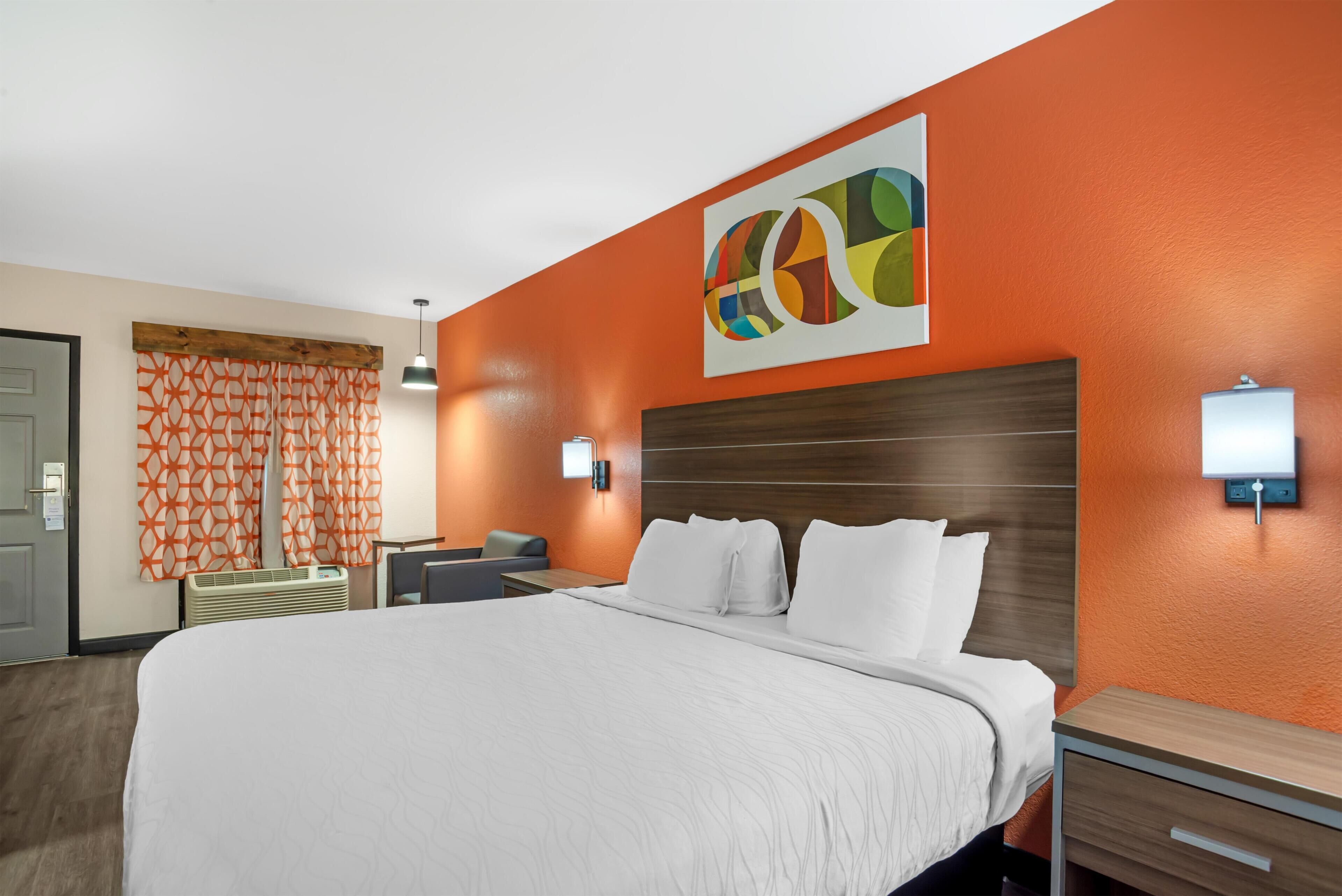 A hotel room with a large bed, a wooden headboard, a wall-mounted light, and a colorful abstract painting.