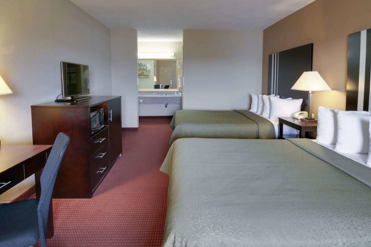 Een hotelkamer met twee bedden, een bureau met stoel, een televisie, een spiegel en een zichtbare badkamer op de achtergrond.