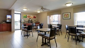 Un comedor amplio con varias mesas y sillas, una barra de cocina con cafeteras y un televisor mostrando un programa.